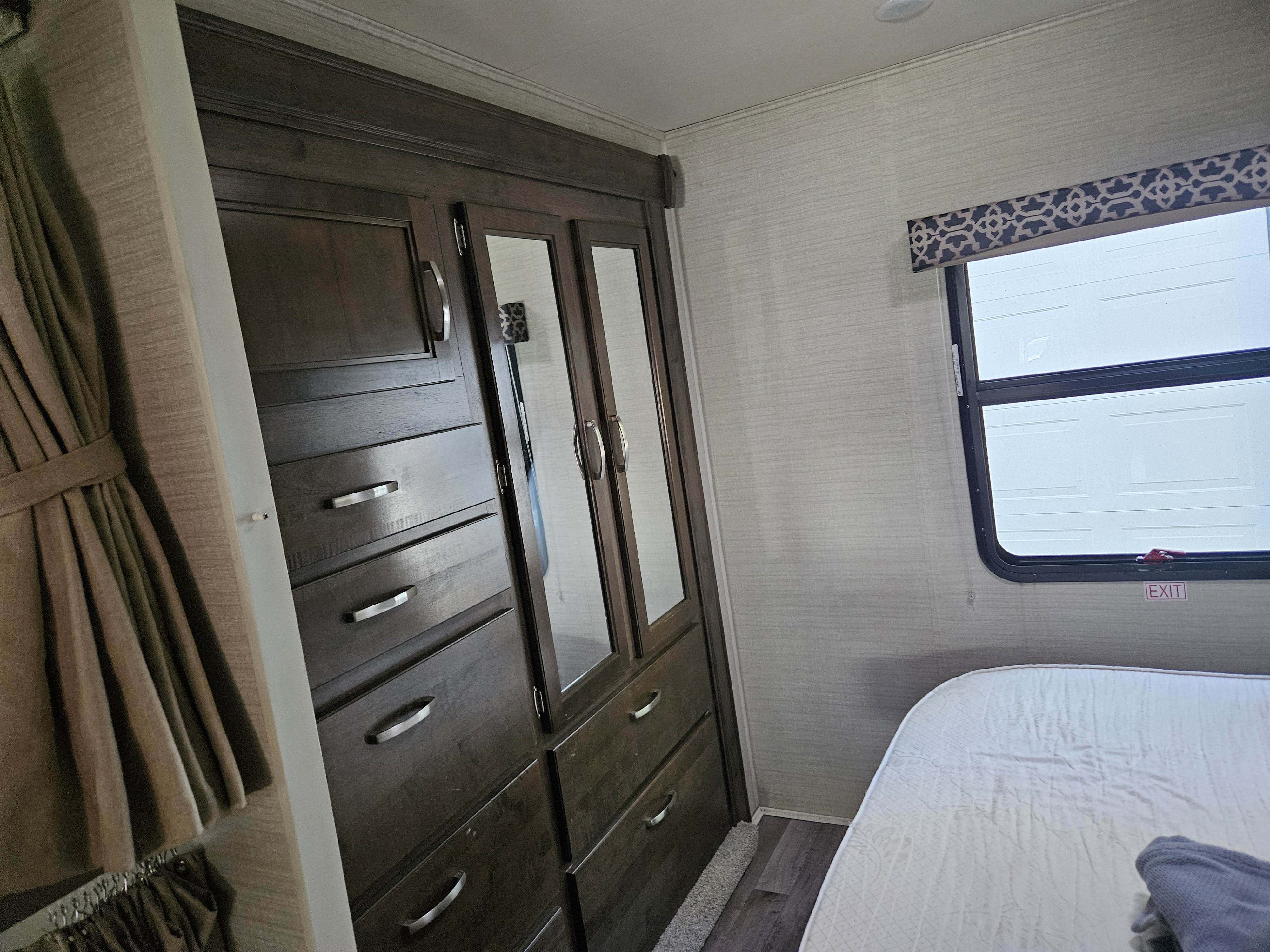 RV rental bedroom with dark wood wardrobe, mirrored doors, and a bed. Shows textured walls and a window with valance.. Jayco Redhawk 2019