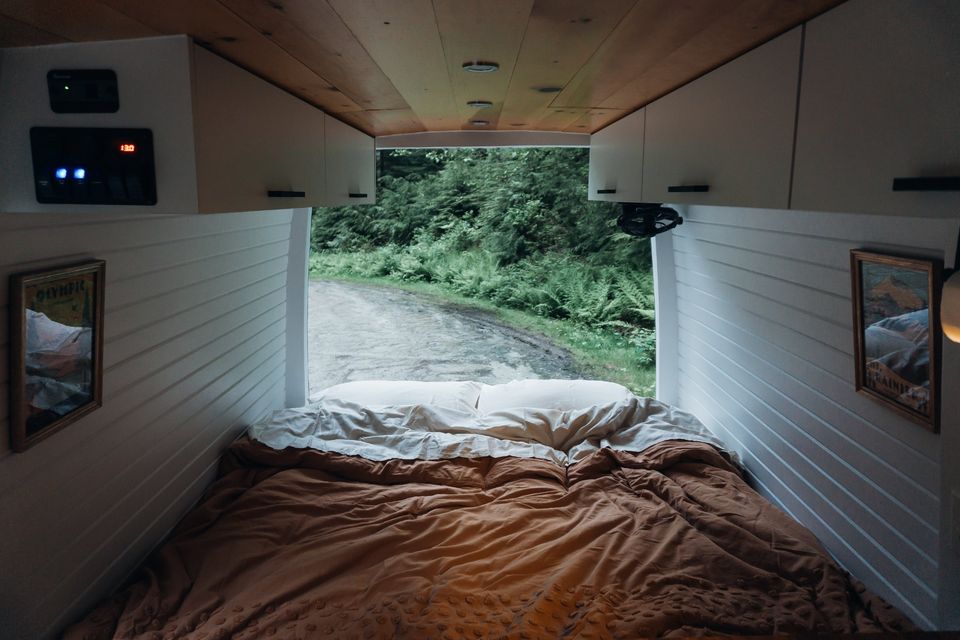 Inside an RV rental, a comfy bed looks out to a forest road. White paneled walls, wood ceiling, controls visible.. Mercedes Benz Sprinter 2500 144 WB 2017
