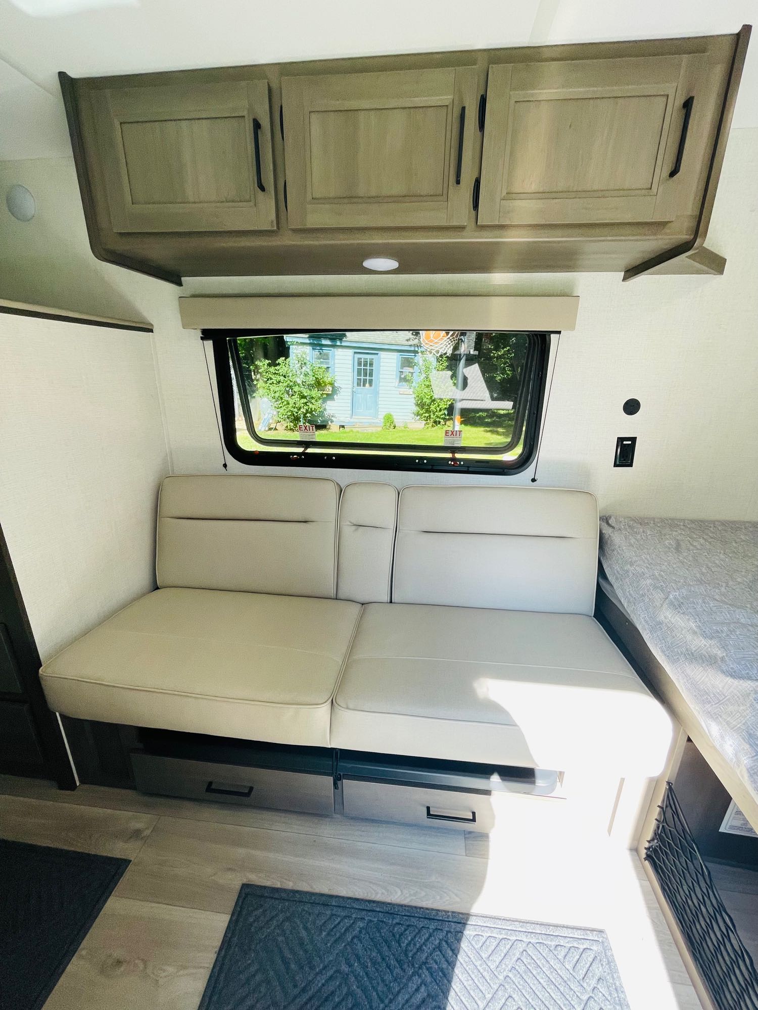 RV rental interior: light sofa, overhead cabinets, window overlooking a house, and bed on the right.. Grand Design Transcend One 151BH 2025