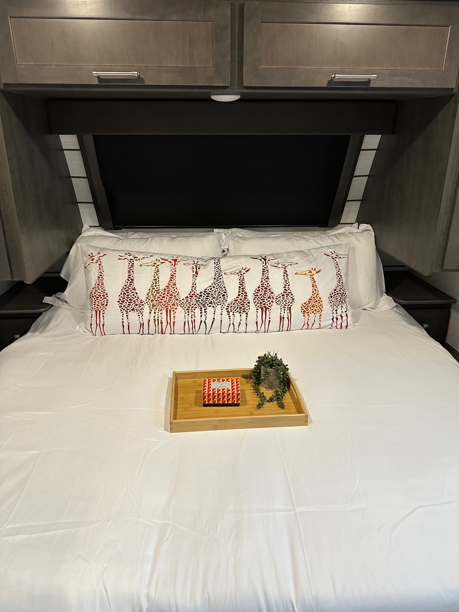A cozy RV rental bedroom shows a bed with white bedding, a giraffe pillow, and dark overhead cabinets.. Keystone RV Cougar 2024
