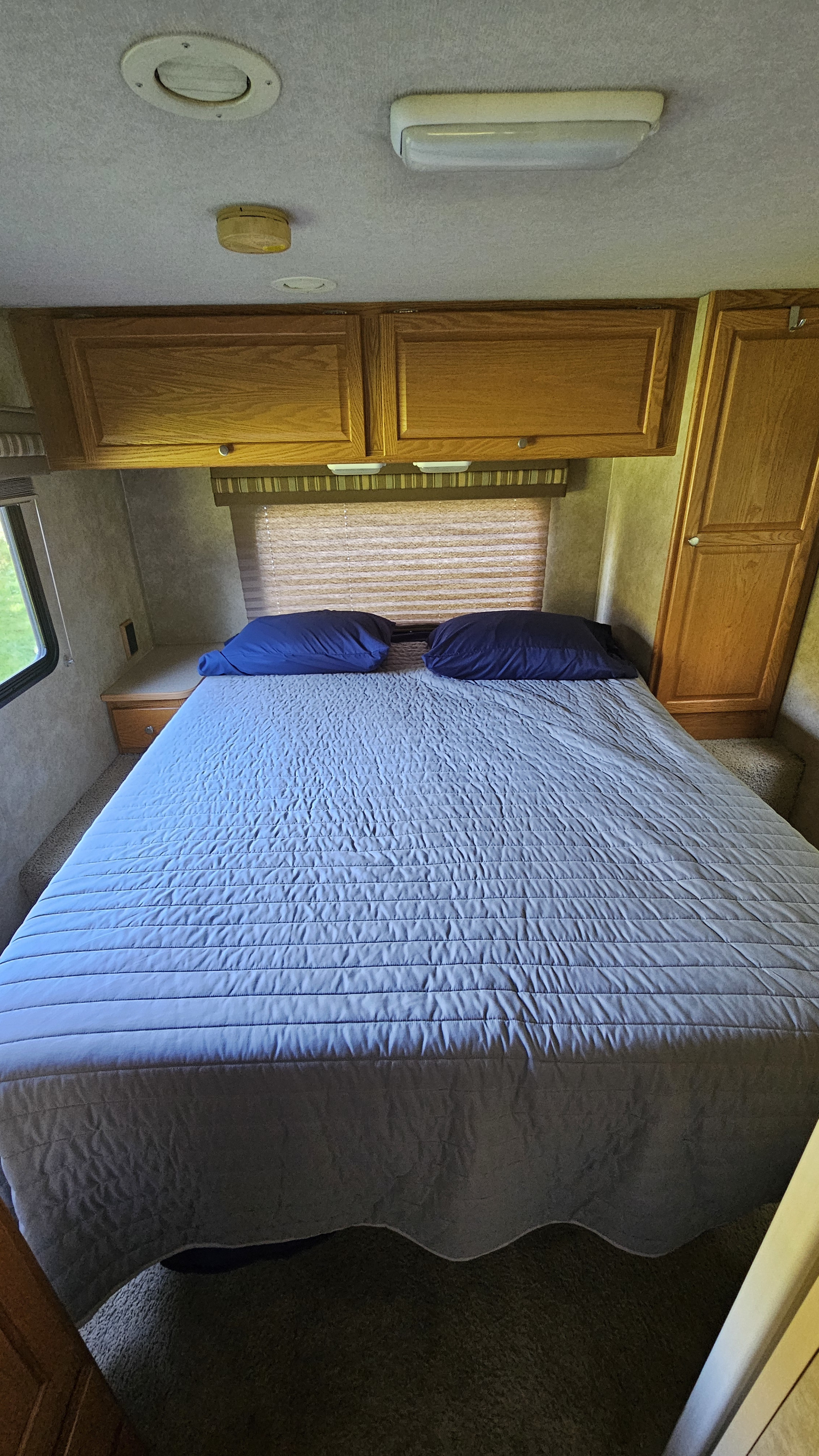 Cozy RV rental bedroom featuring a blue-covered bed, wood overhead cabinets, and a window. Perfect for your travels.. Winnebago Access 2008
