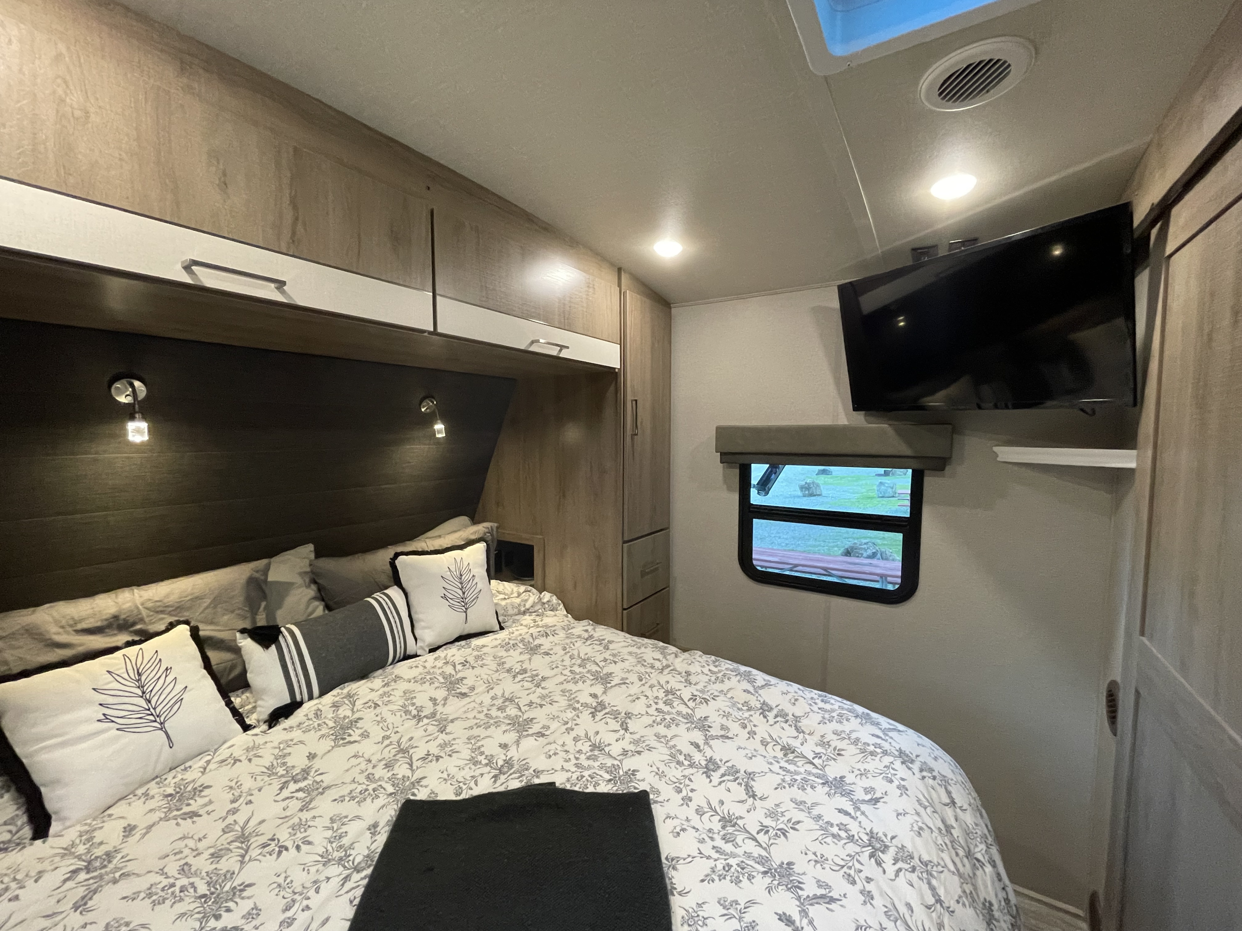 Inside an RV rental: a neat bedroom with floral duvet, mounted TV, storage, and a scenic window view.. Grand Design Imagine 2022