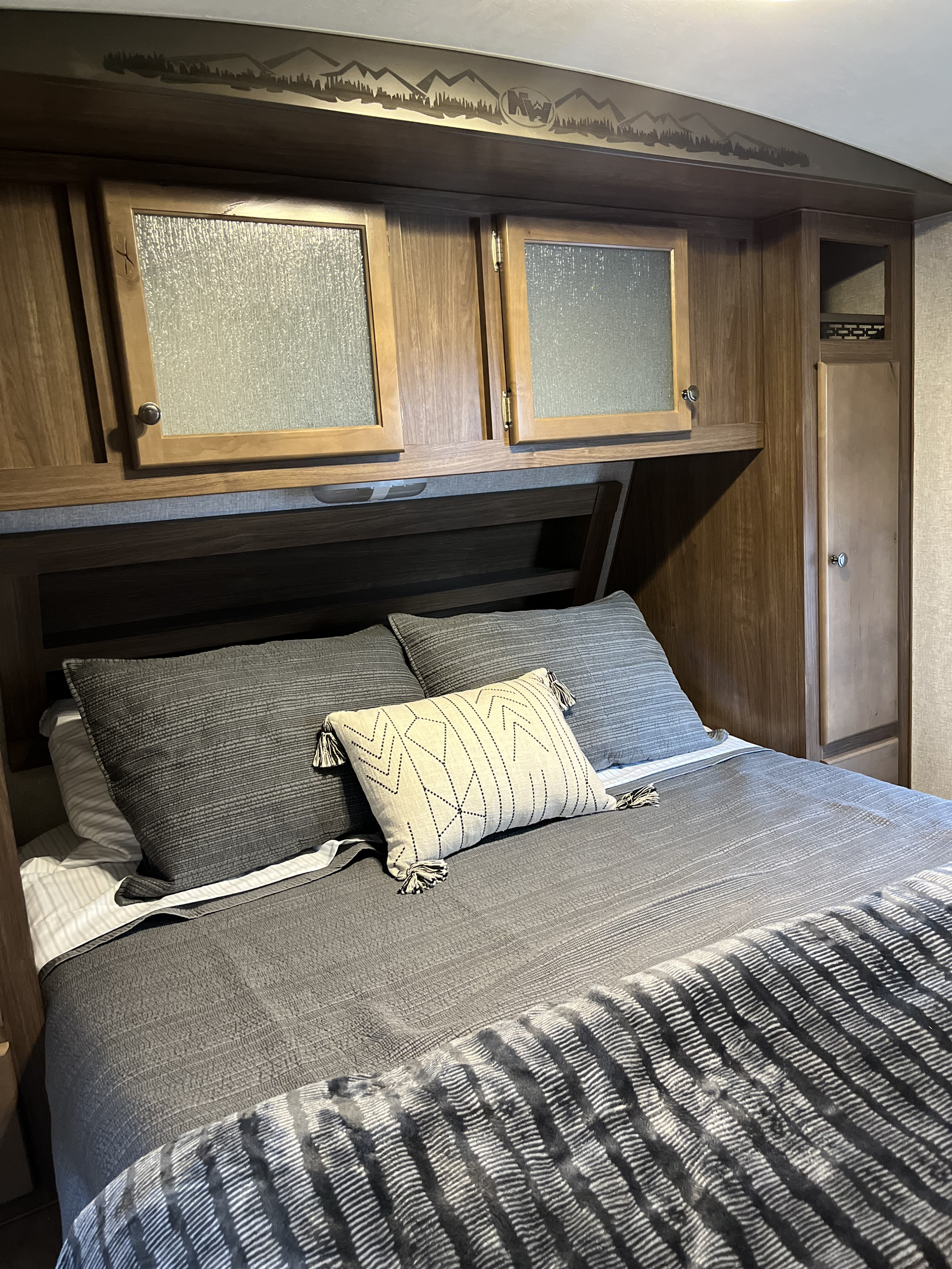 RV rental bedroom: Cozy bed with gray linens, intricate throw pillow, overhead wooden cabinets & mountain-themed decor.. Nash Nash Trailer 2021