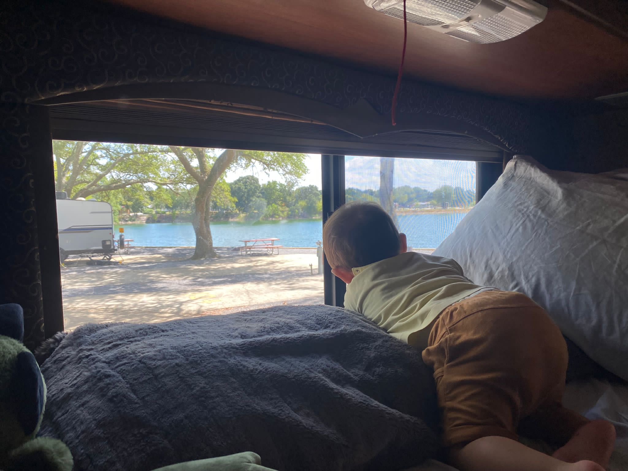 From a cozy RV rental, a baby gazes out at a sunny lake, trees, and another RV. Perfect for family travel & comfort.. Thor Four Winds 2015