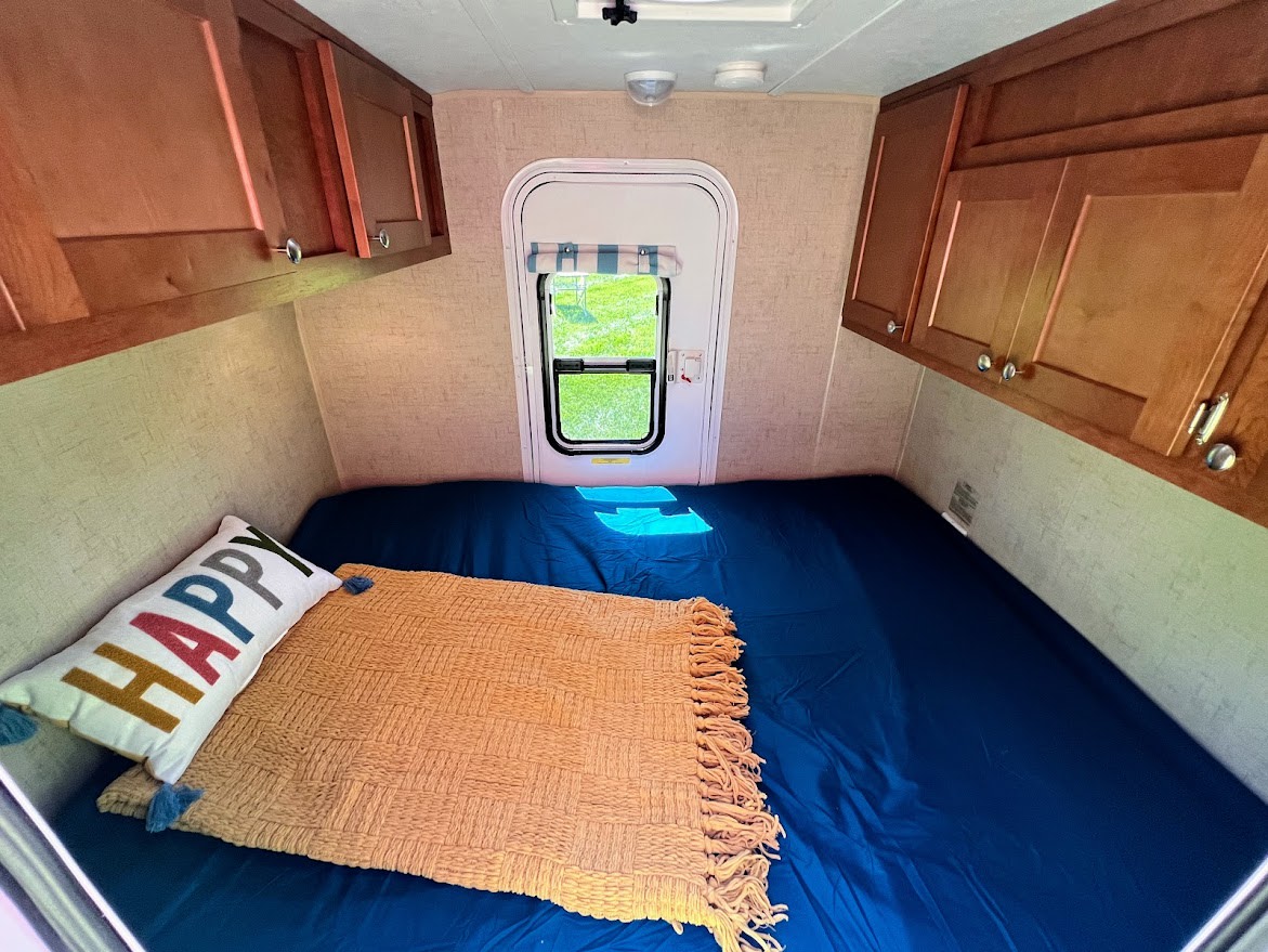 Cozy RV rental interior: bed with 'HAPPY' pillow, ample storage cabinets, and a window view of nature.. Riverside RV White Water 2016