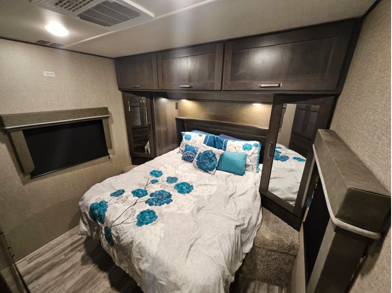 Cozy RV bedroom for your next RV rental. Features a queen bed with blue floral bedding, overhead storage, and a built-in mirror.. Grand Design Reflection 2022