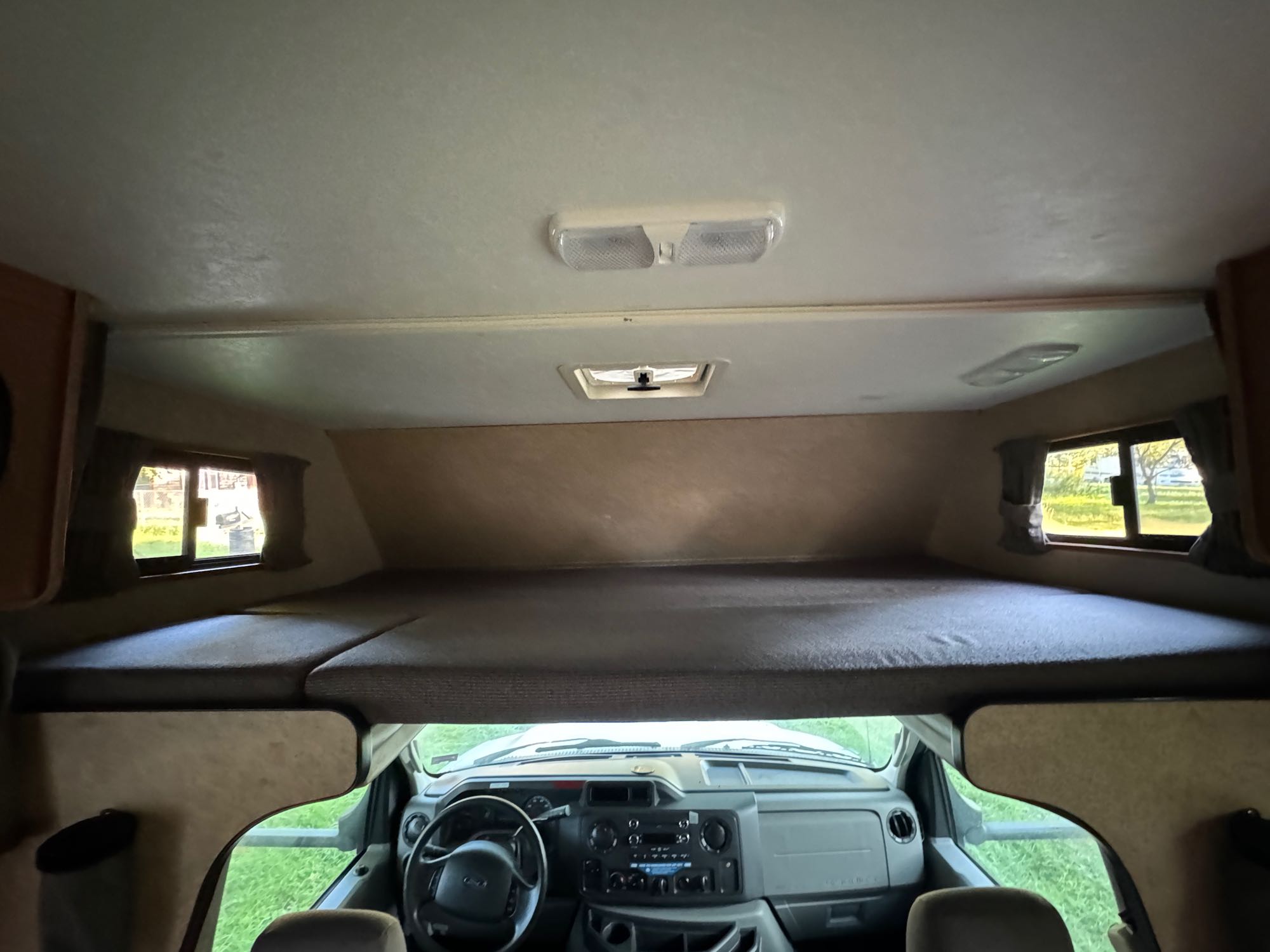 RV rental interior showing a cabover bed above the cockpit with steering wheel. Ceiling lights and a vent are visible.. Thor Majestic 2017