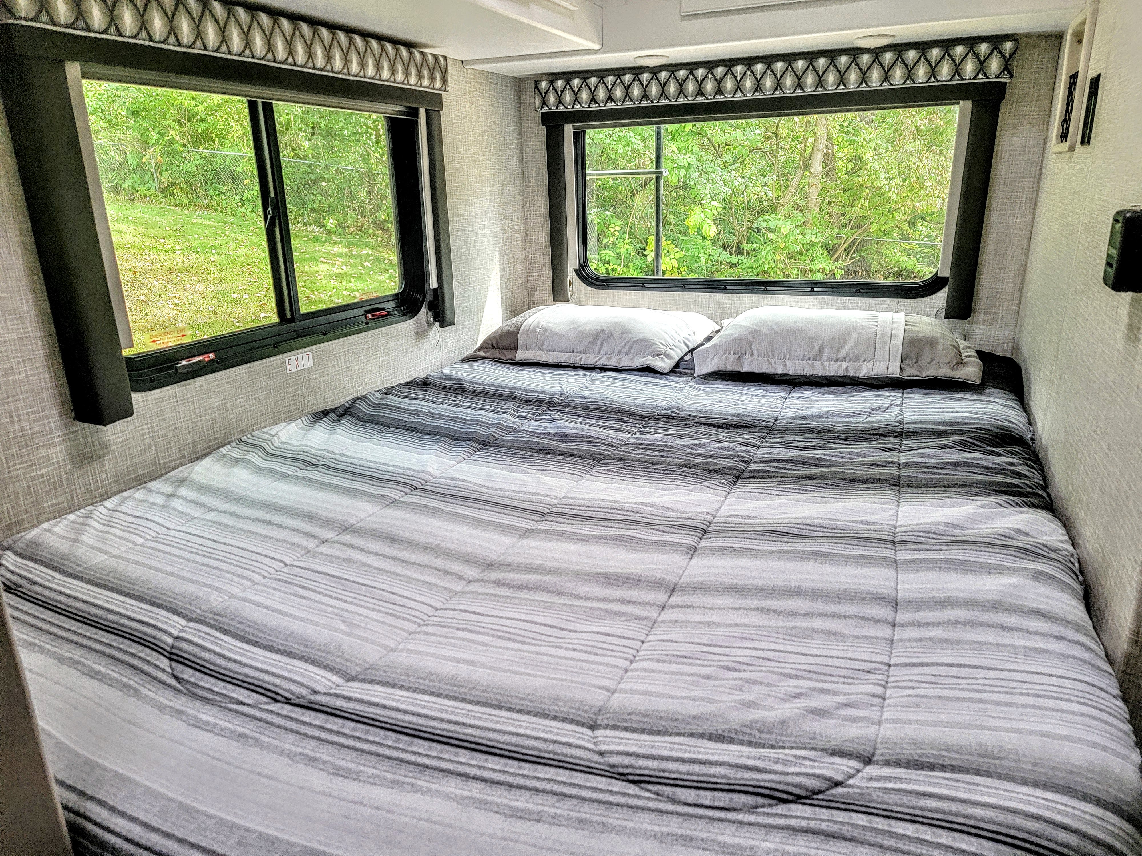 Comfortable RV rental bedroom with a grey striped bed, two pillows, and large windows offering a lush green view.. Thor Geneva 22VT 2025