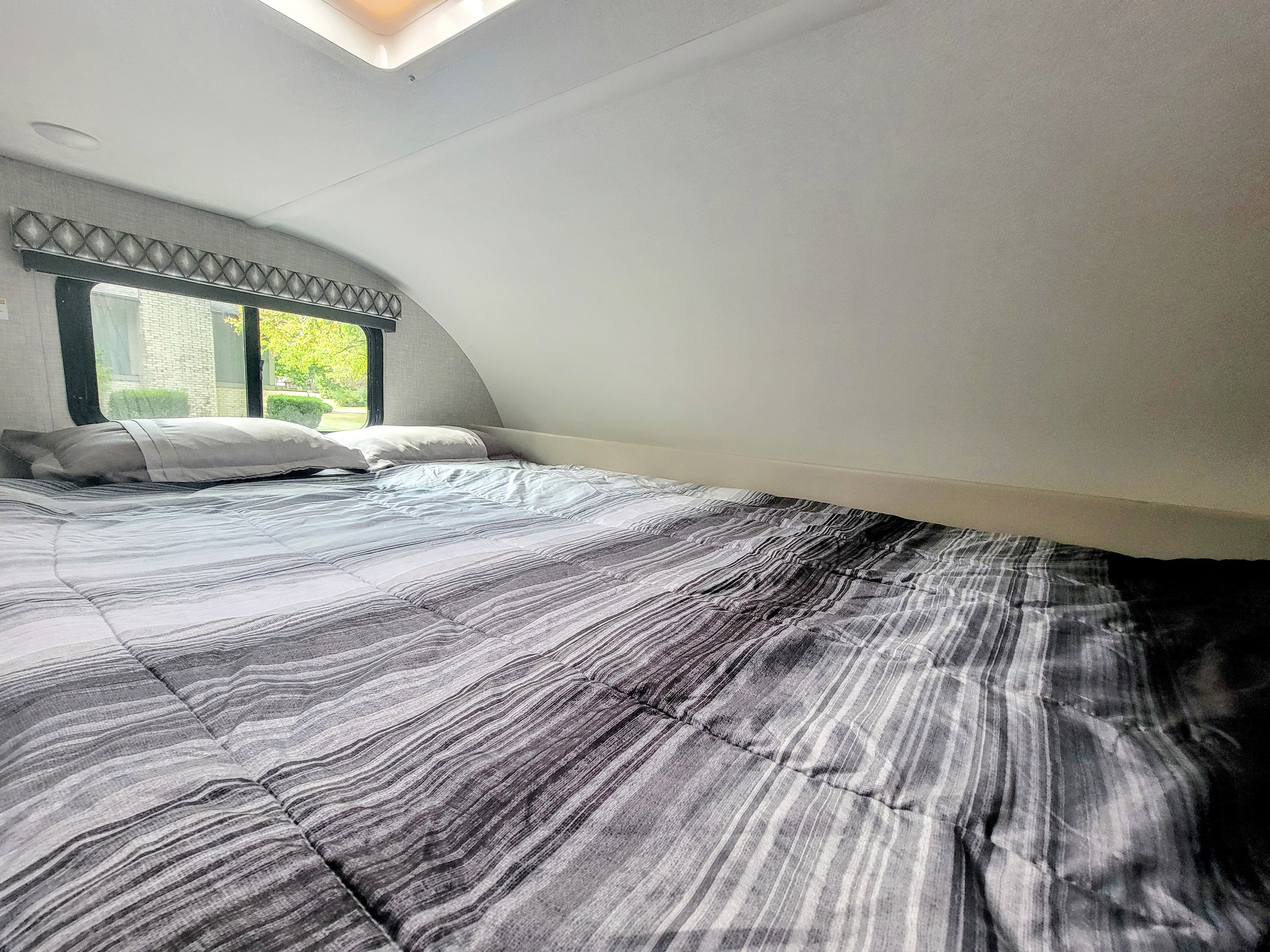 RV rental's cozy cab-over bed, featuring grey striped bedding, pillows, and a bright window view.. Thor Geneva 22VT 2025