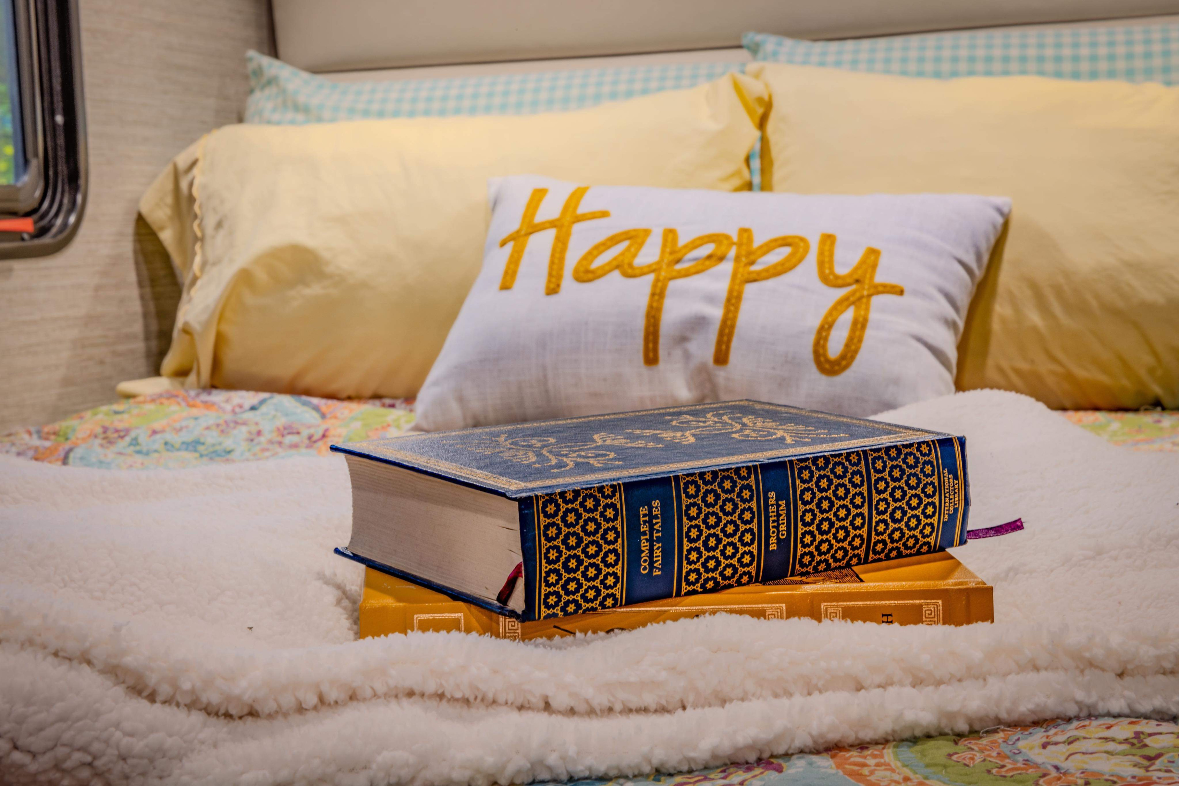 Cozy RV rental bed with a 'Happy' pillow, soft blanket, and stacked books, including Grimm's Fairy Tales.. Winnebago Minnie Winnie 2021