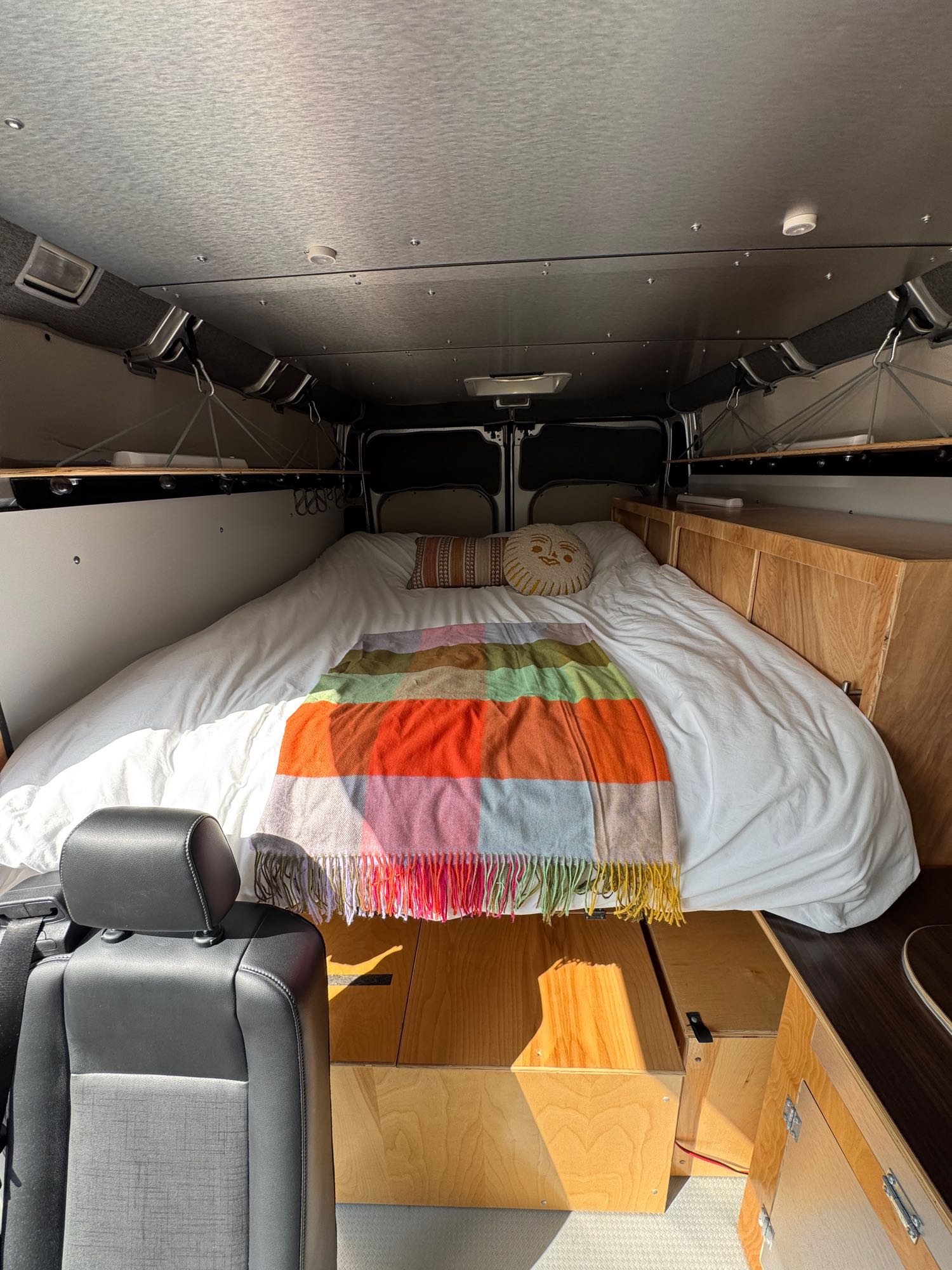Cozy RV rental interior: A spacious bed with a vibrant blanket, surrounded by custom wooden storage and shelving.. Ram Promaster 2500 2021