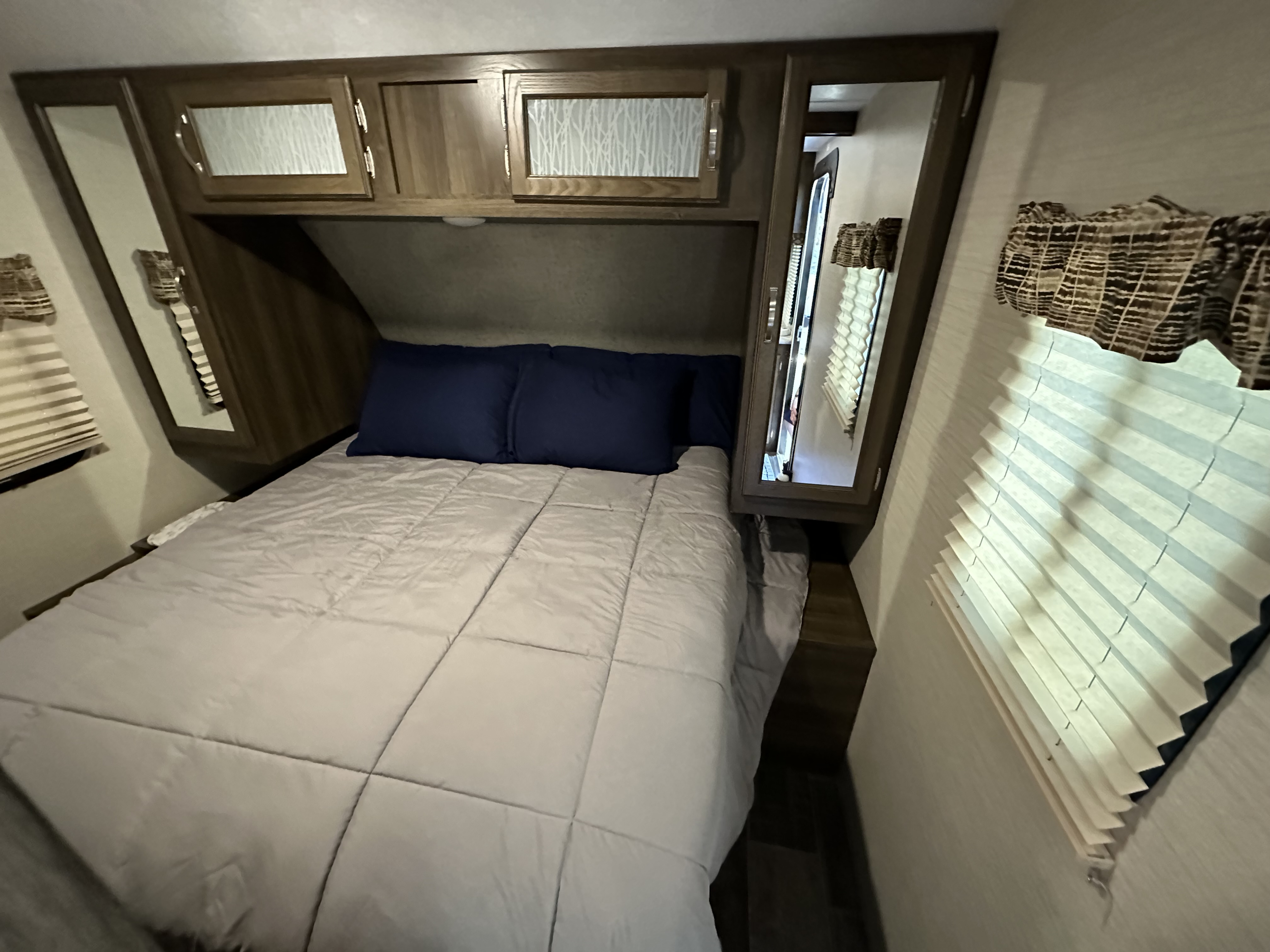 RV rental bedroom: cozy space with bed, blue pillows, overhead cabinets, mirrored doors, and pleated window blinds.. Keystone RV Bullet Ultra Lite 2018
