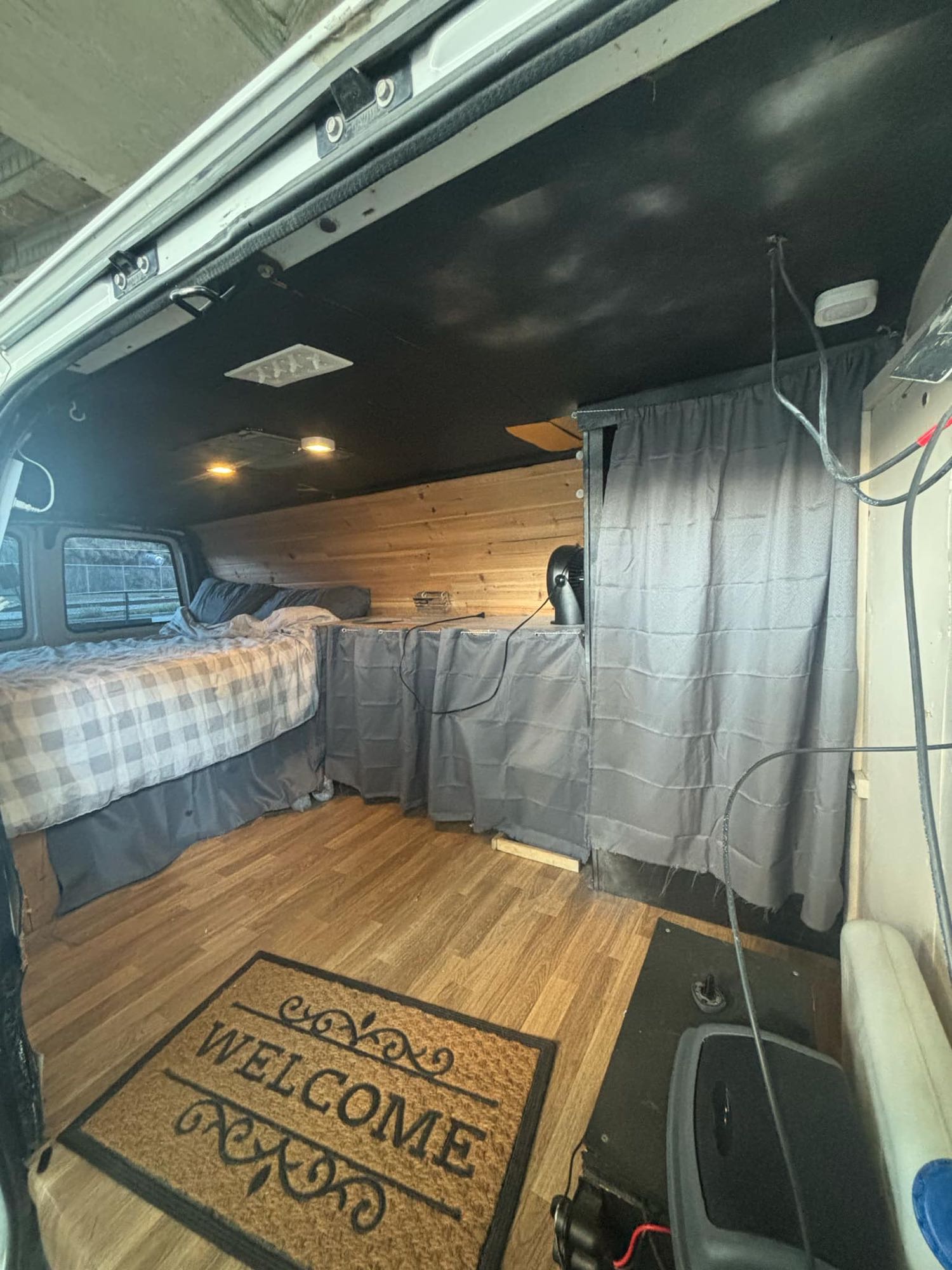 Cozy RV rental camper van interior: a comfortable bed with plaid bedding, wood paneling, and a 'Welcome' mat on the floor.. Gmc Savanna 1998