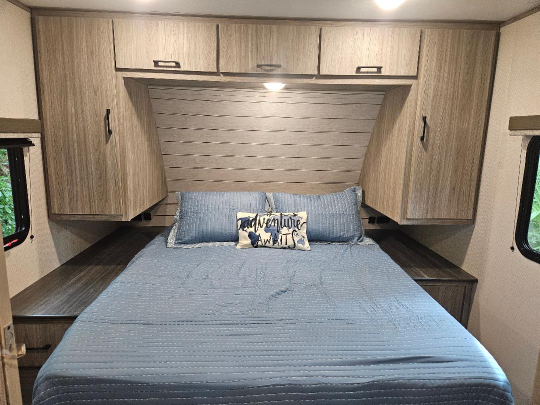 Cozy RV rental bedroom with blue bedding, overhead cabinets, and a scenic window view. Adventure awaits!. Keystone RV Passport SL 2021