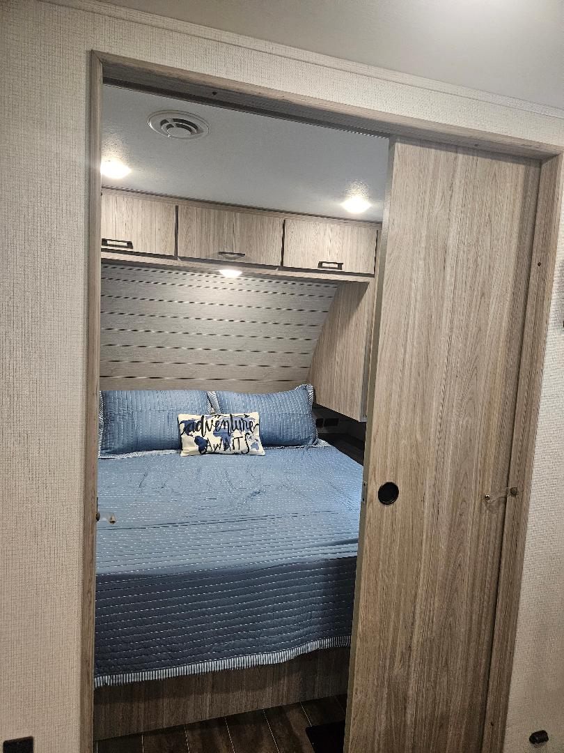 Inside an RV rental: a cozy bedroom with a blue bed, "Adventure Awaits" pillow, overhead cabinets, and wood sliding door.. Keystone RV Passport SL 2021