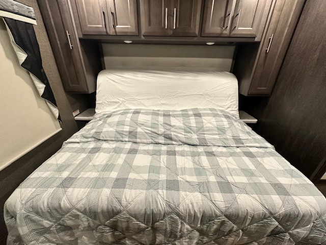 Compact RV bedroom interior shows a bed with a plaid comforter, white sheet, and overhead cabinets. Ideal for RV rental.. Thor Challenger 2022