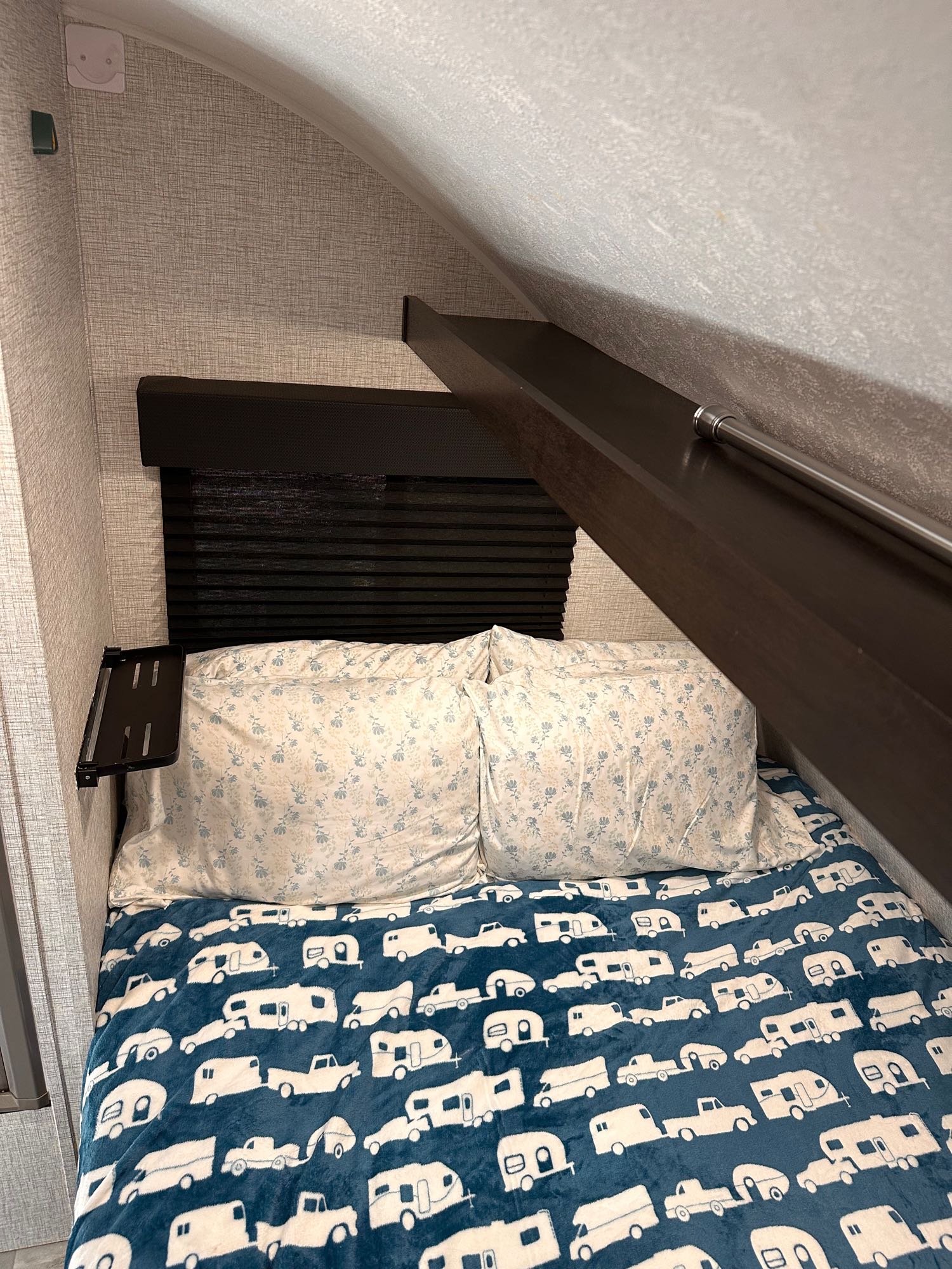 Cozy RV rental bedroom: bed with floral pillows, blue camper blanket, small shelf, and angled ceiling above.. Braxton Creek FreeSolo DIN 2023