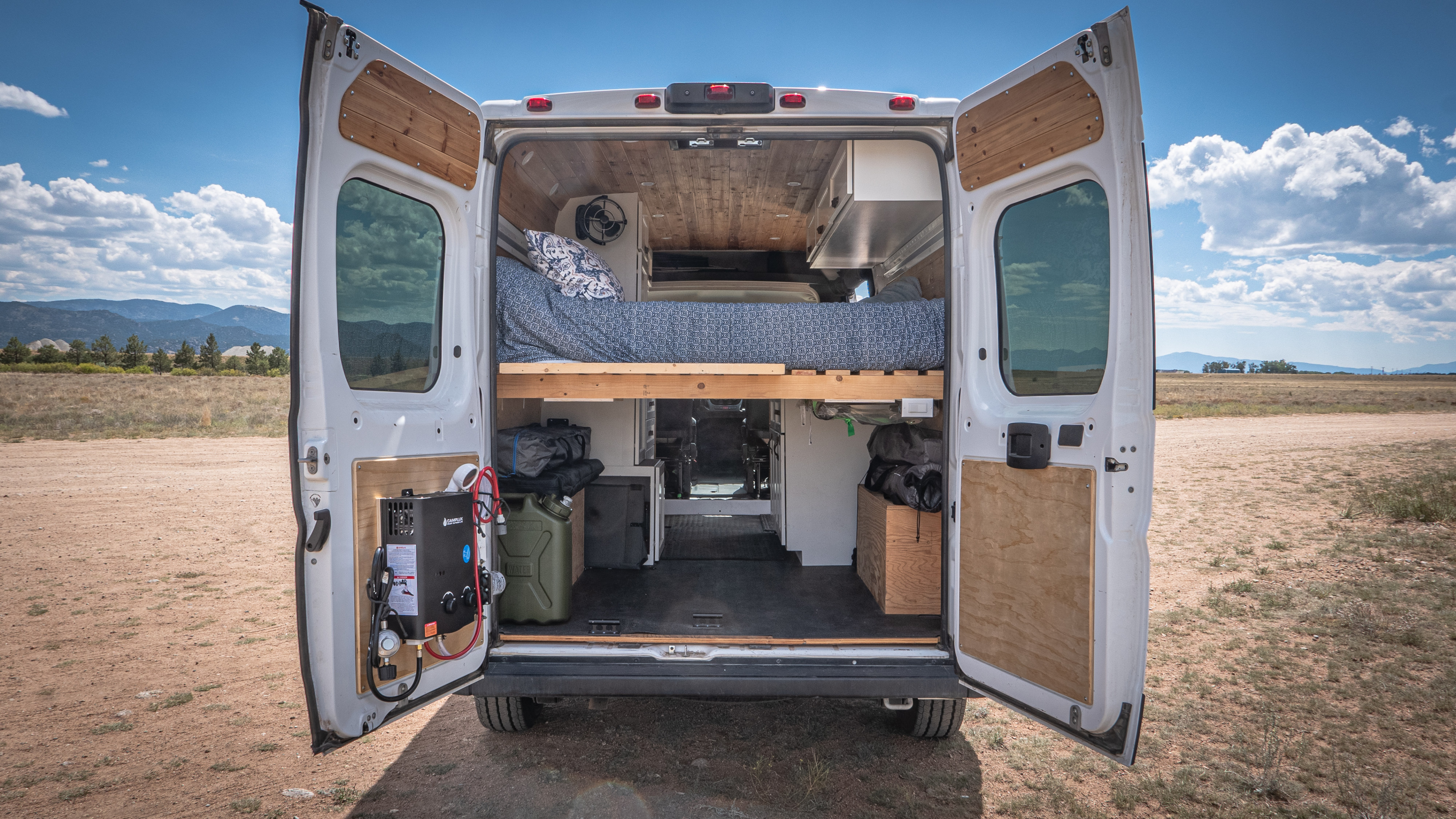 Open back doors of an RV rental van revealing a cozy bed, storage, and a mounted portable water heater, perfect for adventures.. Dodge Promaster 2500 2021