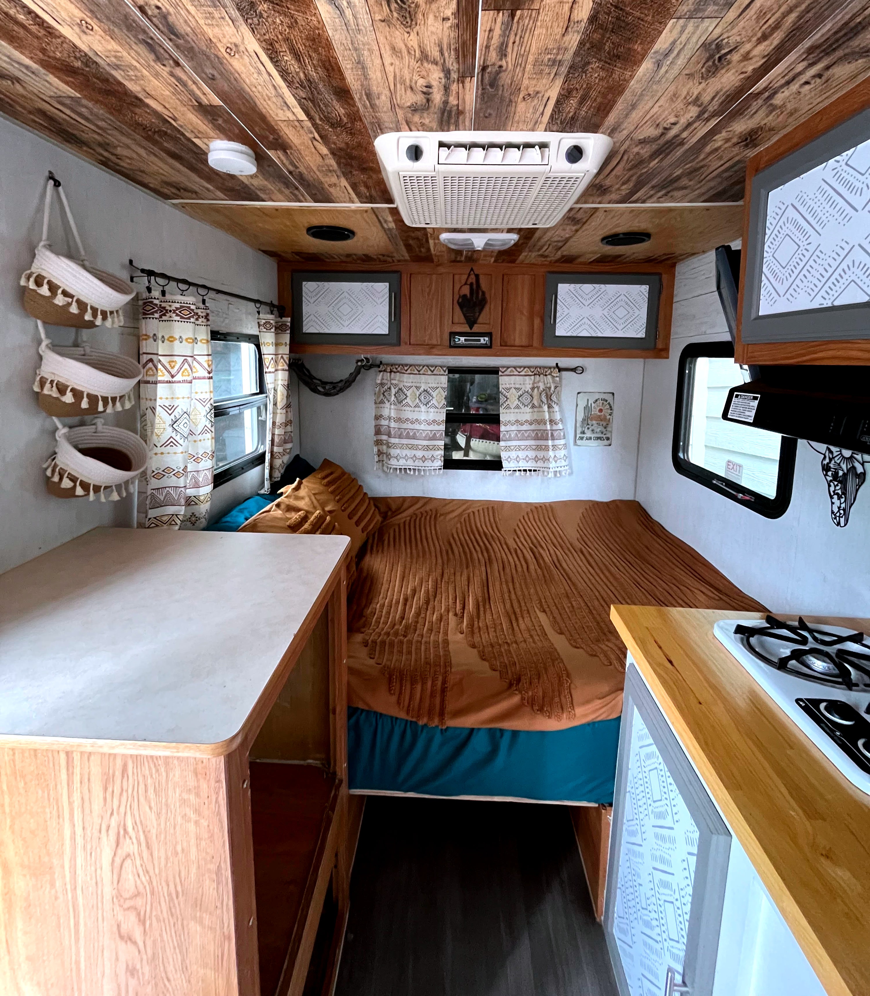 Cozy RV rental interior with a wooden ceiling, terracotta and teal bedding on the bed, and a compact kitchen area.. Sidekick 15F60 2008
