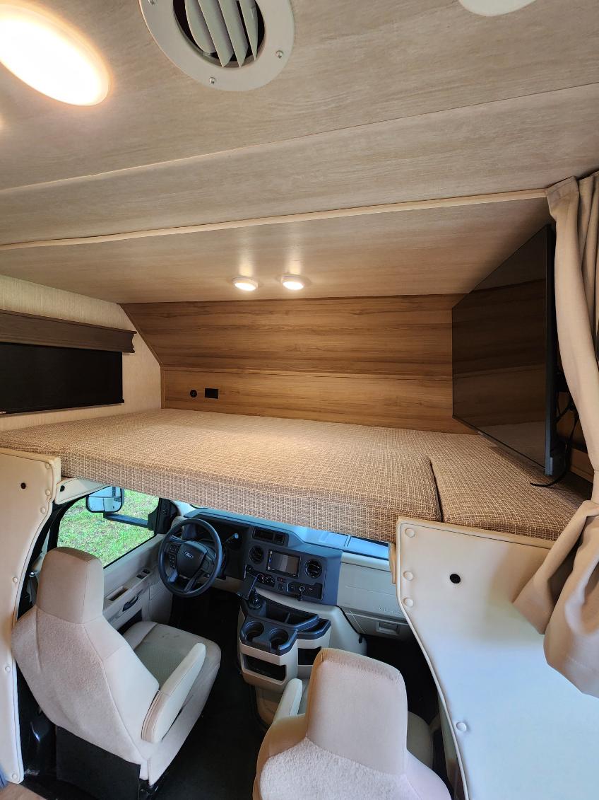 RV rental interior: Front cabin with driver's seat below a spacious overhead bunk bed, featuring a mounted TV.. NeXus Triumph 31T 2024