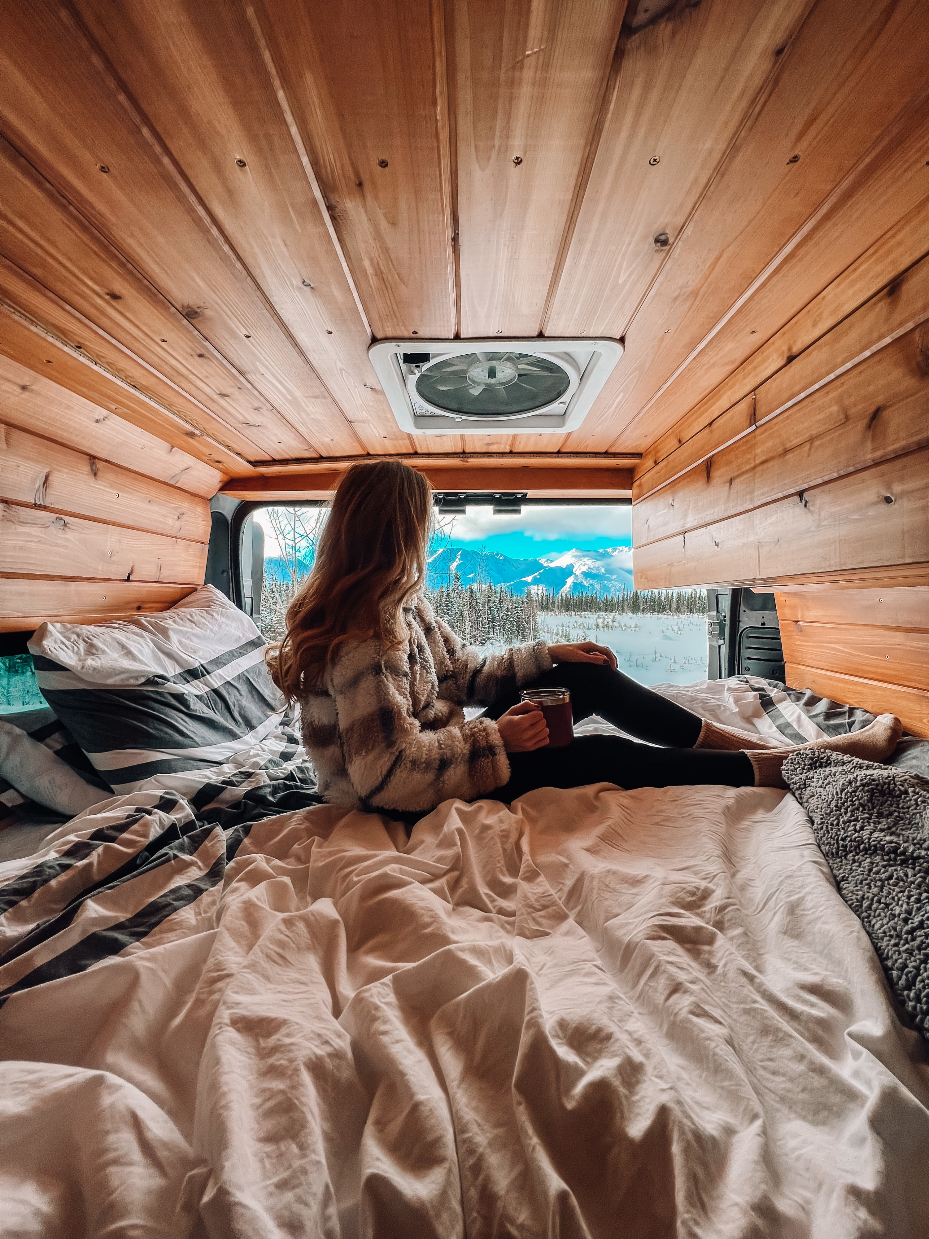 Cozy RV rental interior: A person on a comfy bed sips a drink, enjoying breathtaking snowy mountain views.. Dodge Van 2024