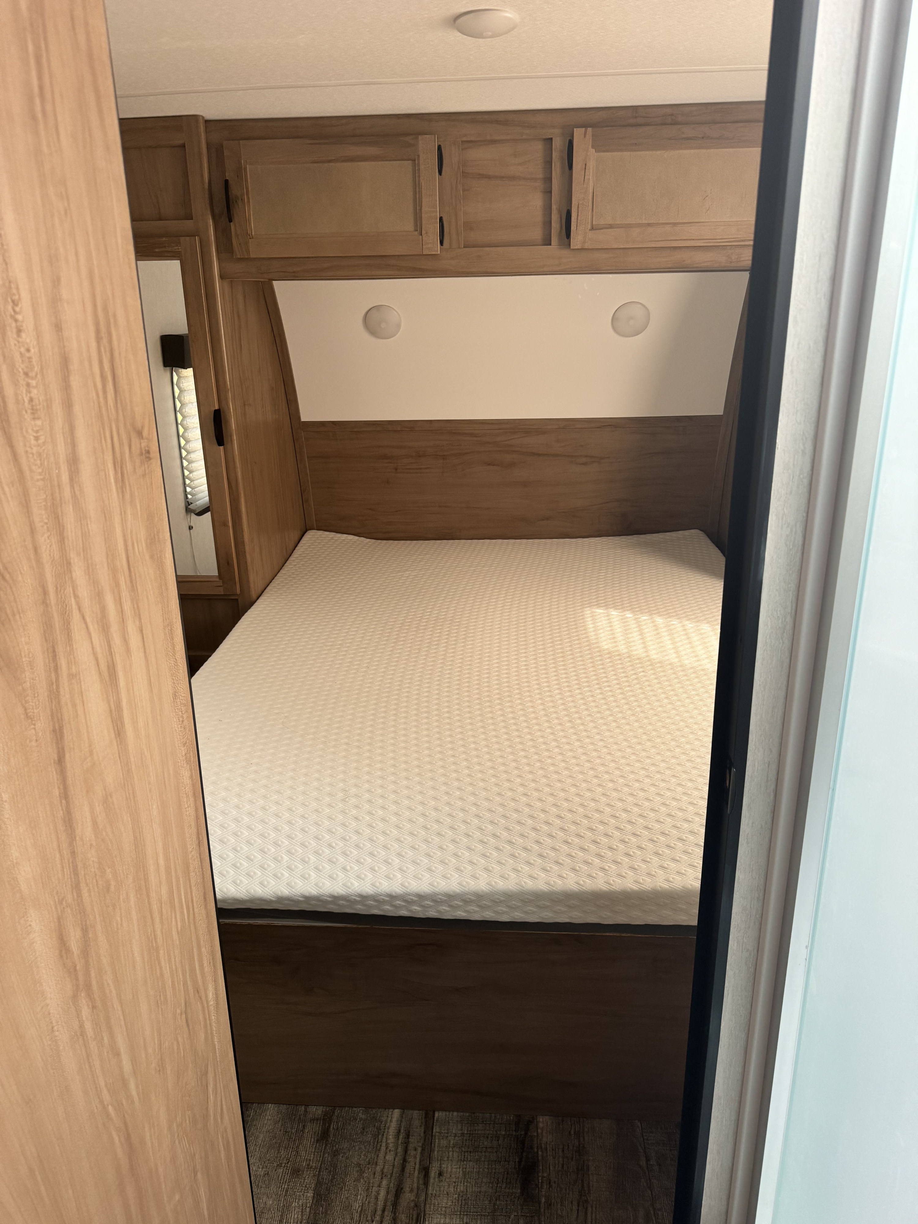 Cozy RV rental bedroom with a white textured mattress, overhead wooden cabinets, and a small mirror on the left.. Forest River 23MK 2024
