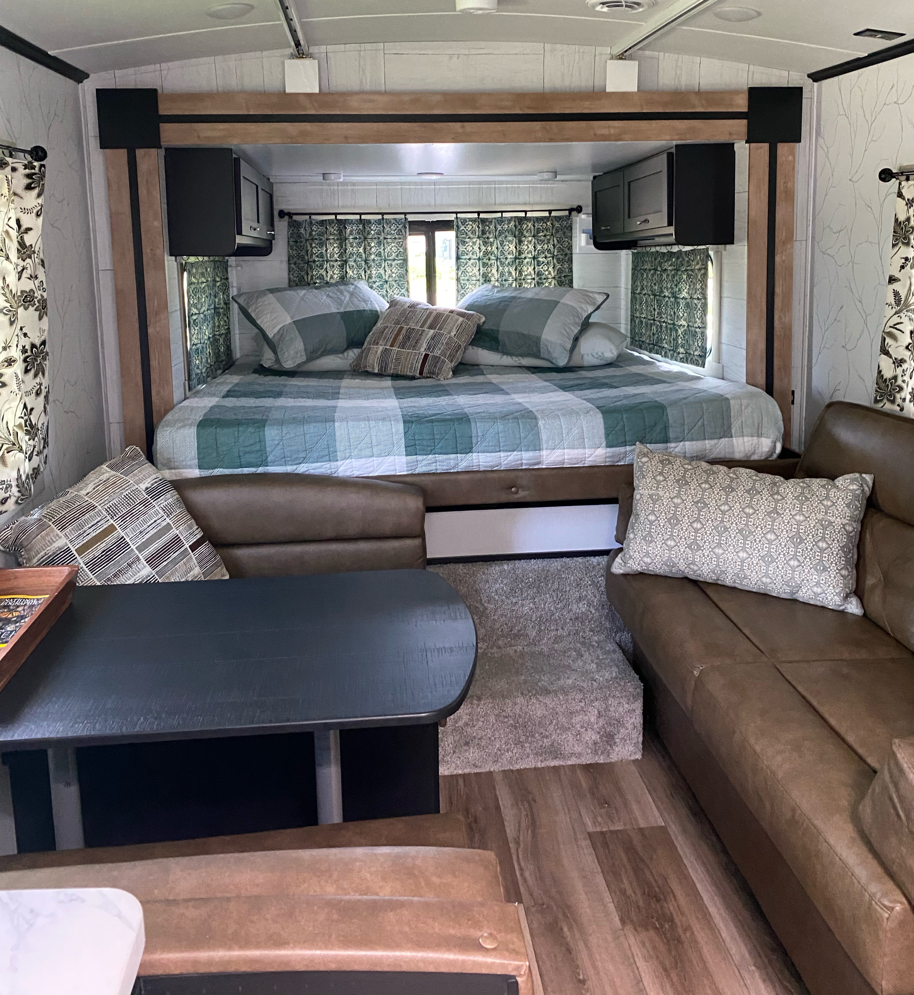 Cozy RV rental interior featuring a comfortable bed with plaid bedding, a brown sofa, and a dark dinette table.. Keystone Outback 2022