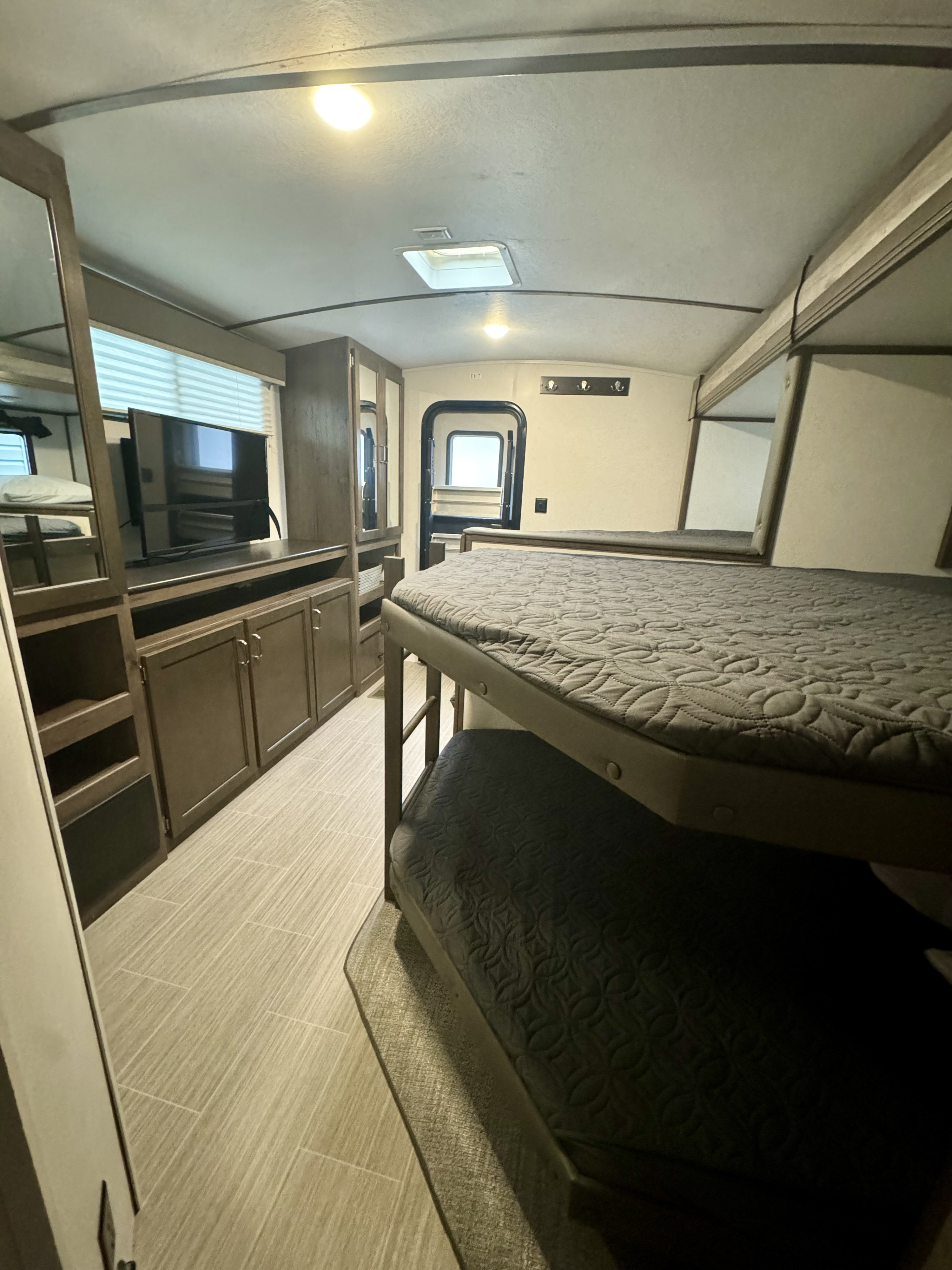 Inside this RV rental, find bunk beds, an entertainment unit with a TV, and an exit door. Bright lights illuminate the interior space.. Keystone RV Passport GT 2021