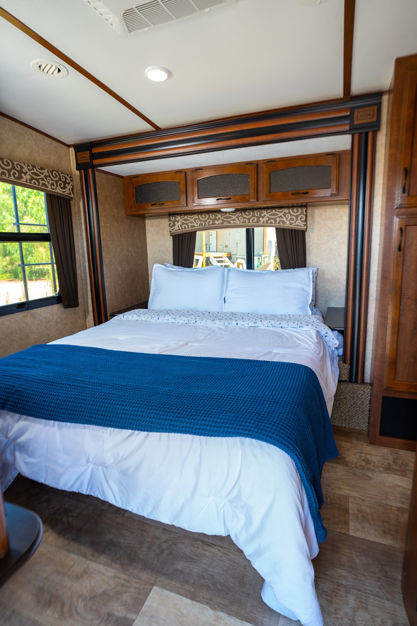 Inside an RV rental bedroom: a neatly made bed with a blue blanket, wood cabinets, and a window view.. Jayco White Hawk 2016