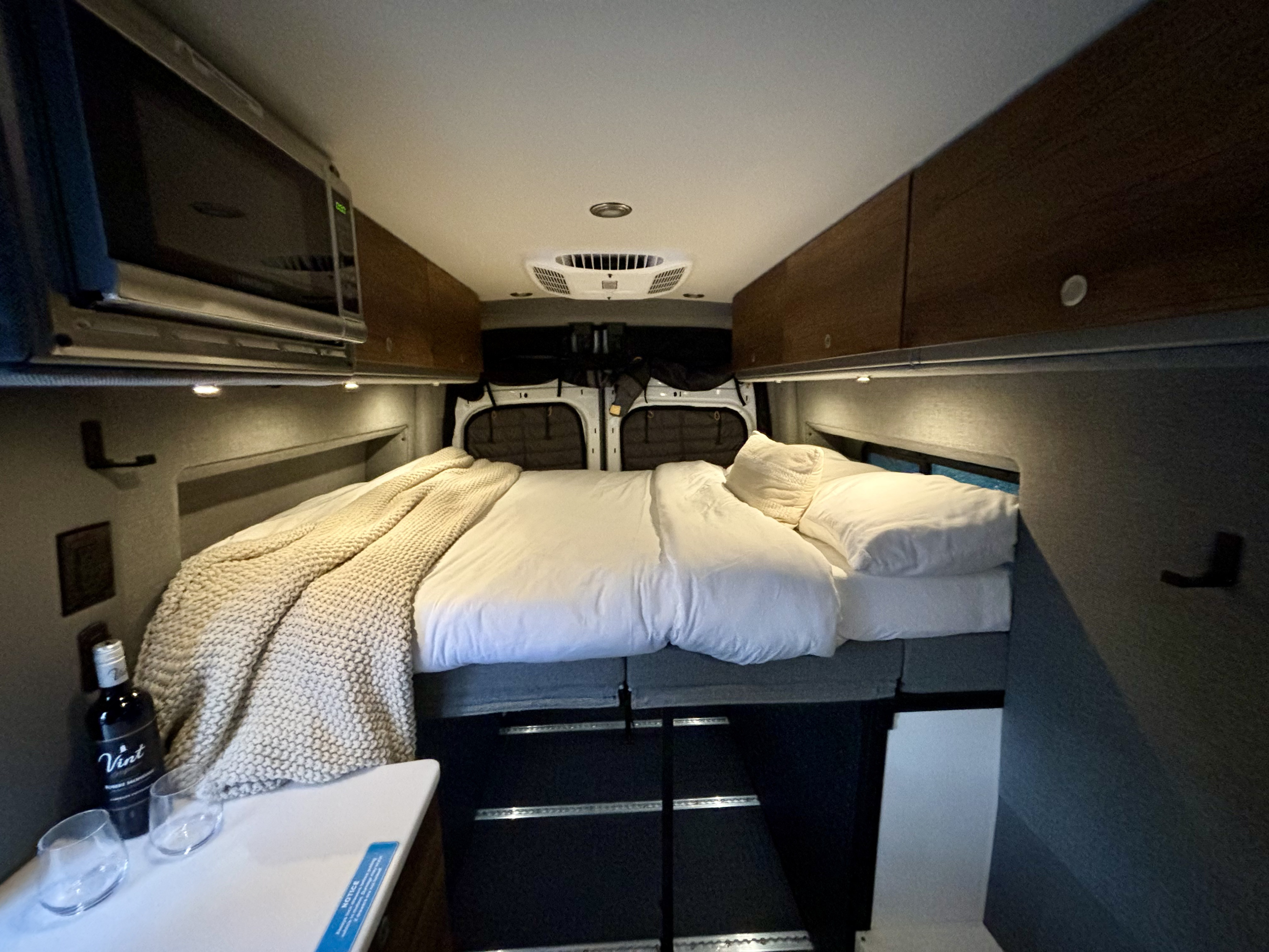 This RV rental features a cozy bed with white linens, overhead storage, a microwave, and wine, offering a comfortable travel interior.. Storyteller Overland Storyteller Overland Class B 2023