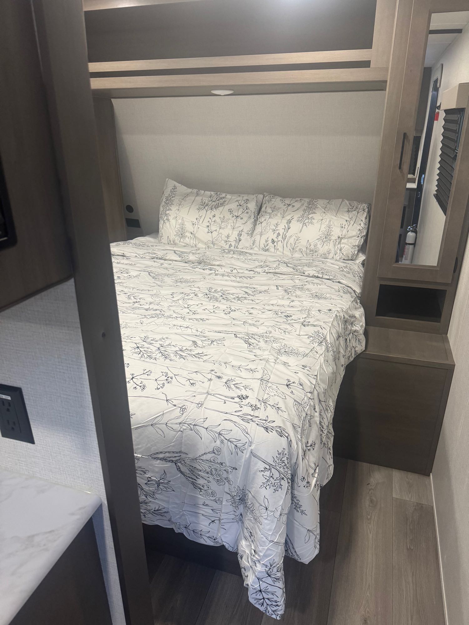 Cozy RV rental bedroom featuring a bed with white floral linens, built-in storage, and wood-look flooring.. Grand Design Transcend Xplor 2024