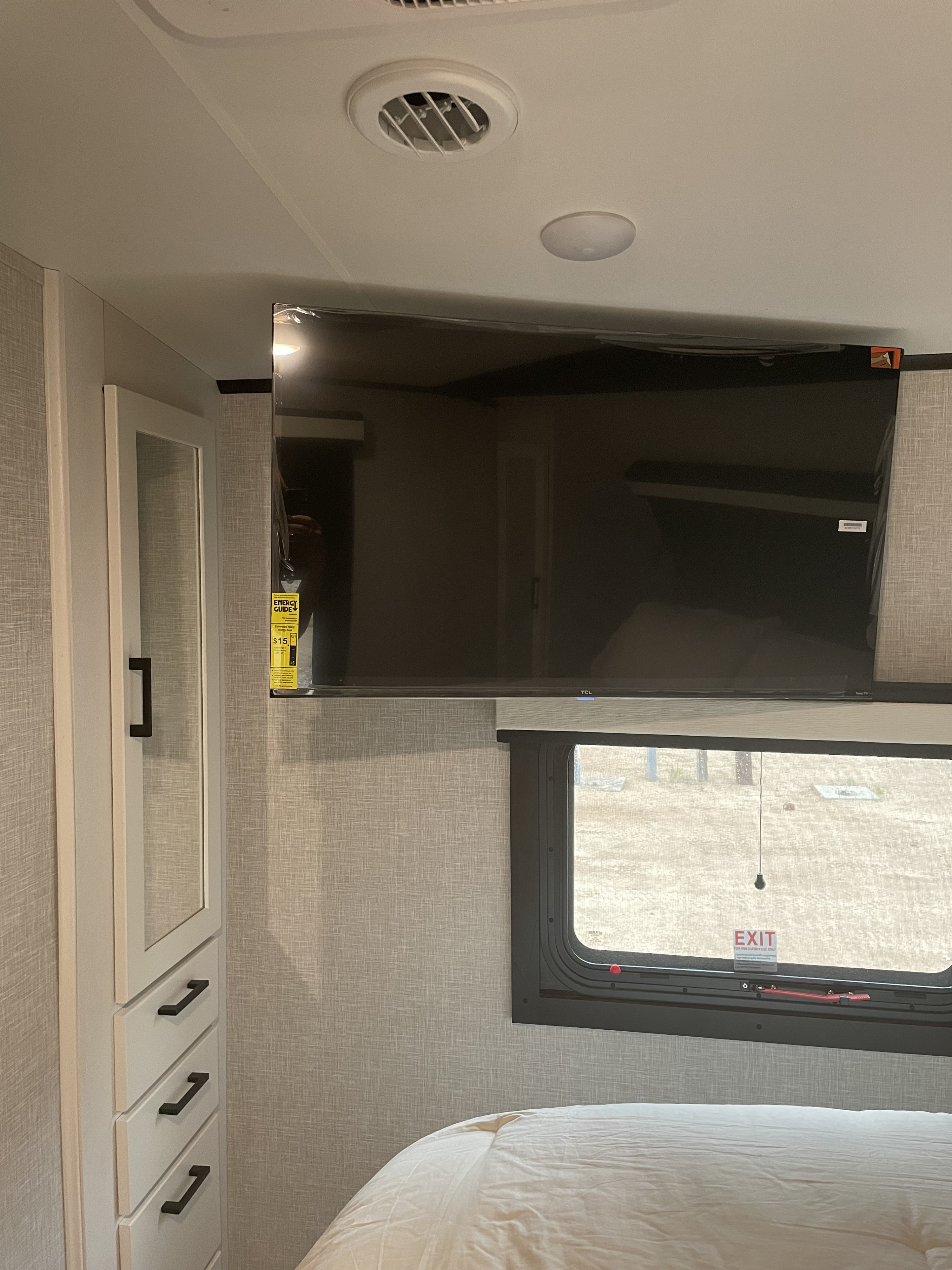 Cozy RV rental bedroom: Features a mounted TV, window, bed, and white storage cabinet. Textured grey walls throughout.. Jayco White Hawk 2024