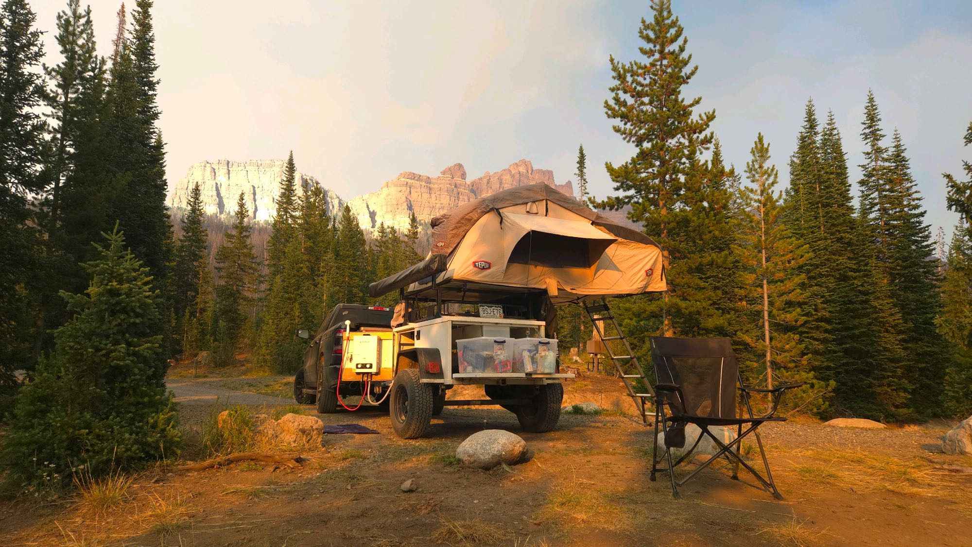 Golden hour camping: truck with rooftop tent and trailer at a forest campsite, mountains behind. An RV rental alternative for adventure.. Other Other 2020