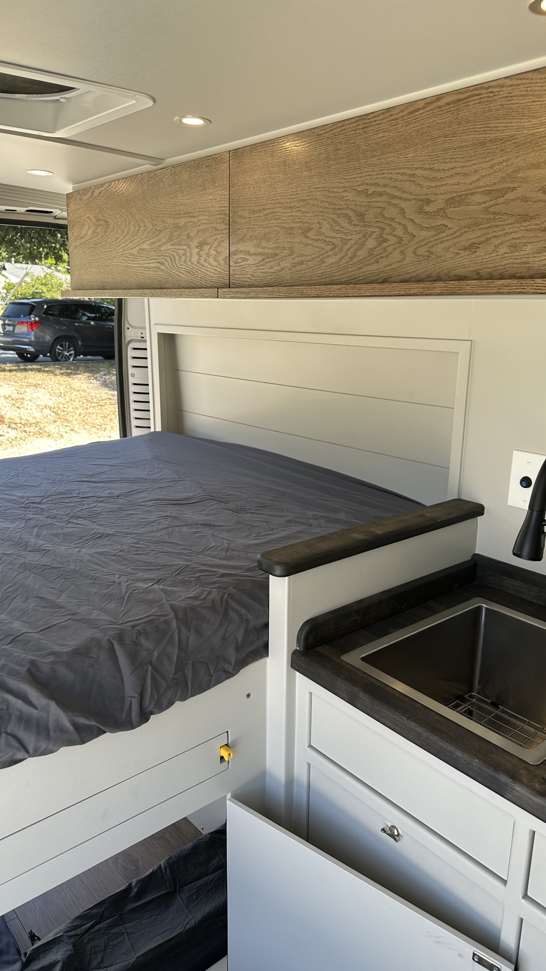 Interior of an rv rental: a grey bed on left, light wood cabinets above, and a compact kitchen sink on the right.. RAM Promaster 2024