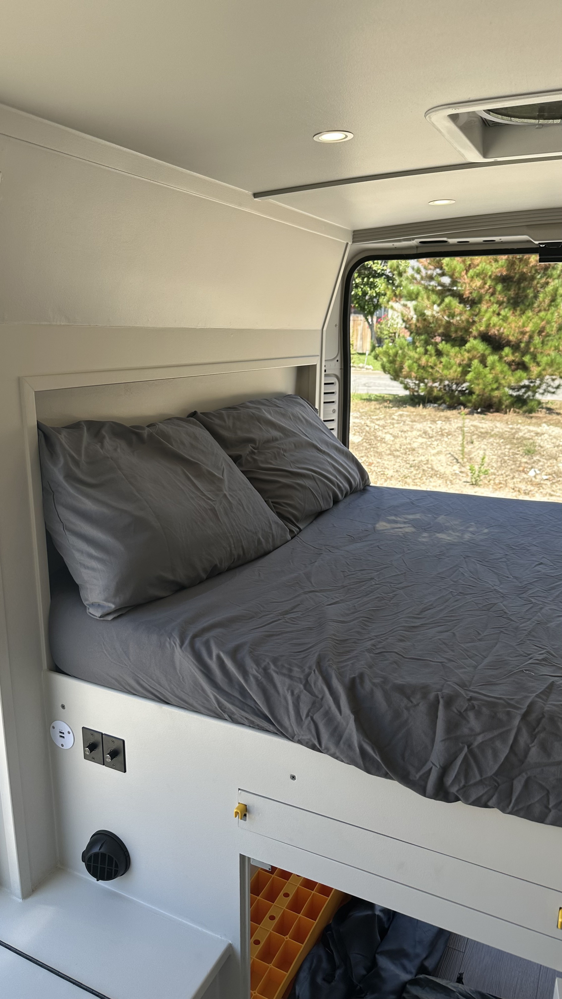A comfortable bed with dark gray bedding and integrated charging ports inside a bright RV rental. Scenic outdoor view.. RAM Promaster 2024