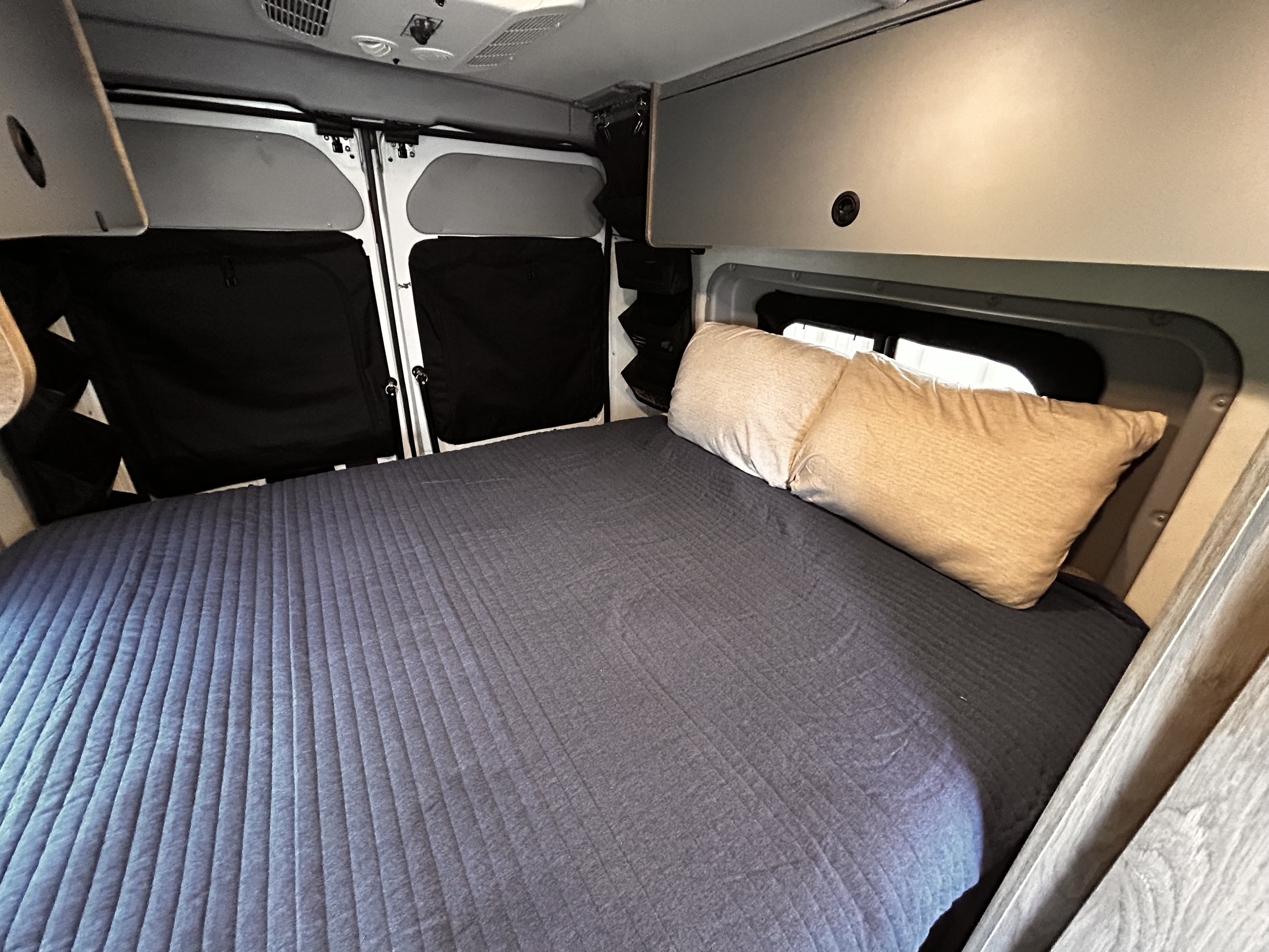 This RV rental's interior features a cozy bed with pillows, window coverings, and overhead storage.. Winnebago Solis 2021