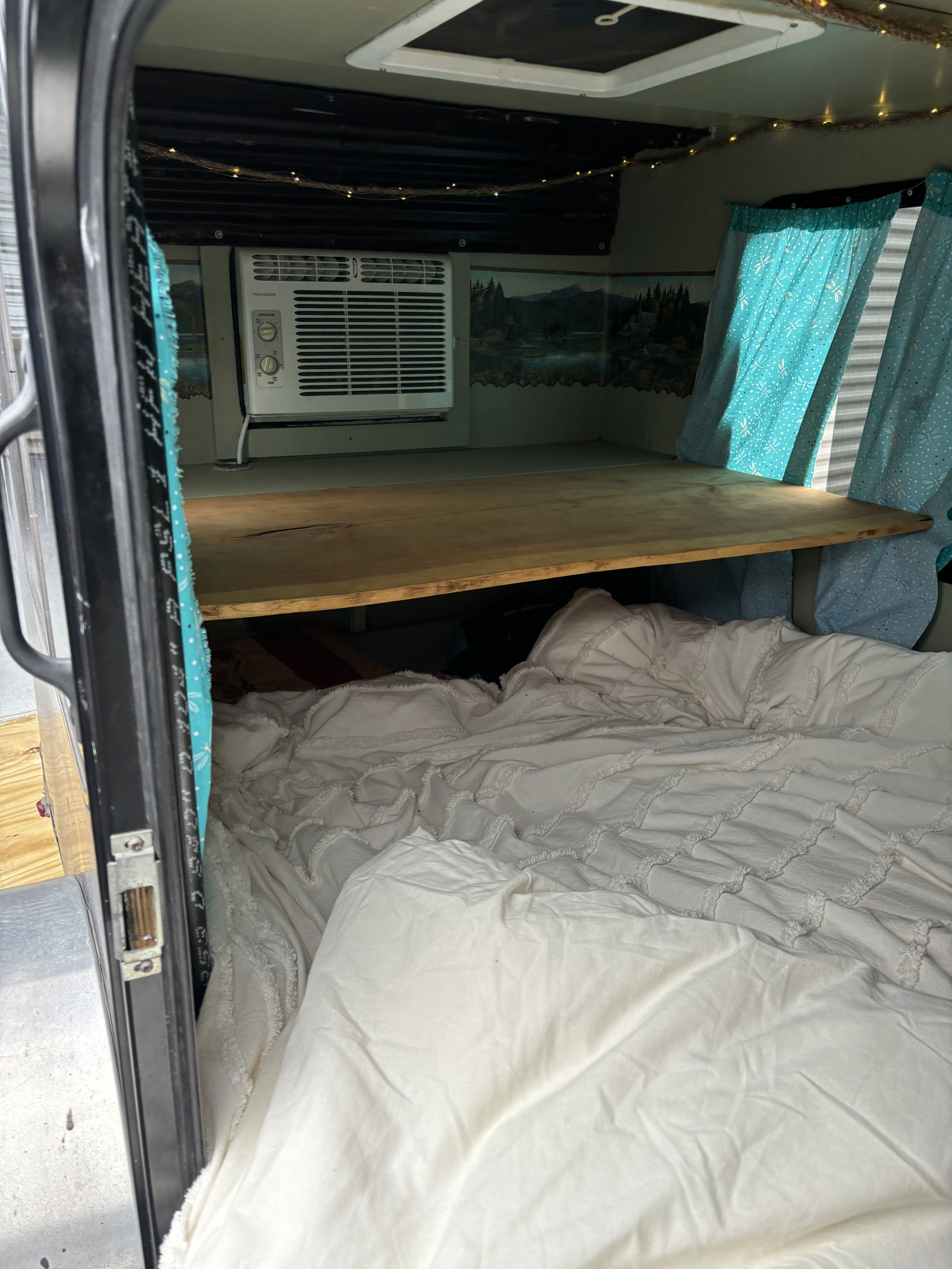 RV rental interior: A cozy bed with white bedding under a wooden shelf, an AC unit, string lights, and a nature mural.. Signatory Tote+4 2014