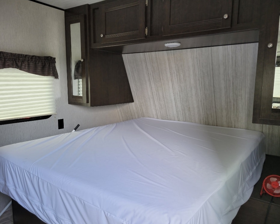 Clean and organized RV rental bedroom featuring a white-covered bed, overhead cabinets, and a window with blinds.. Heartland RVs Pioneer 2019