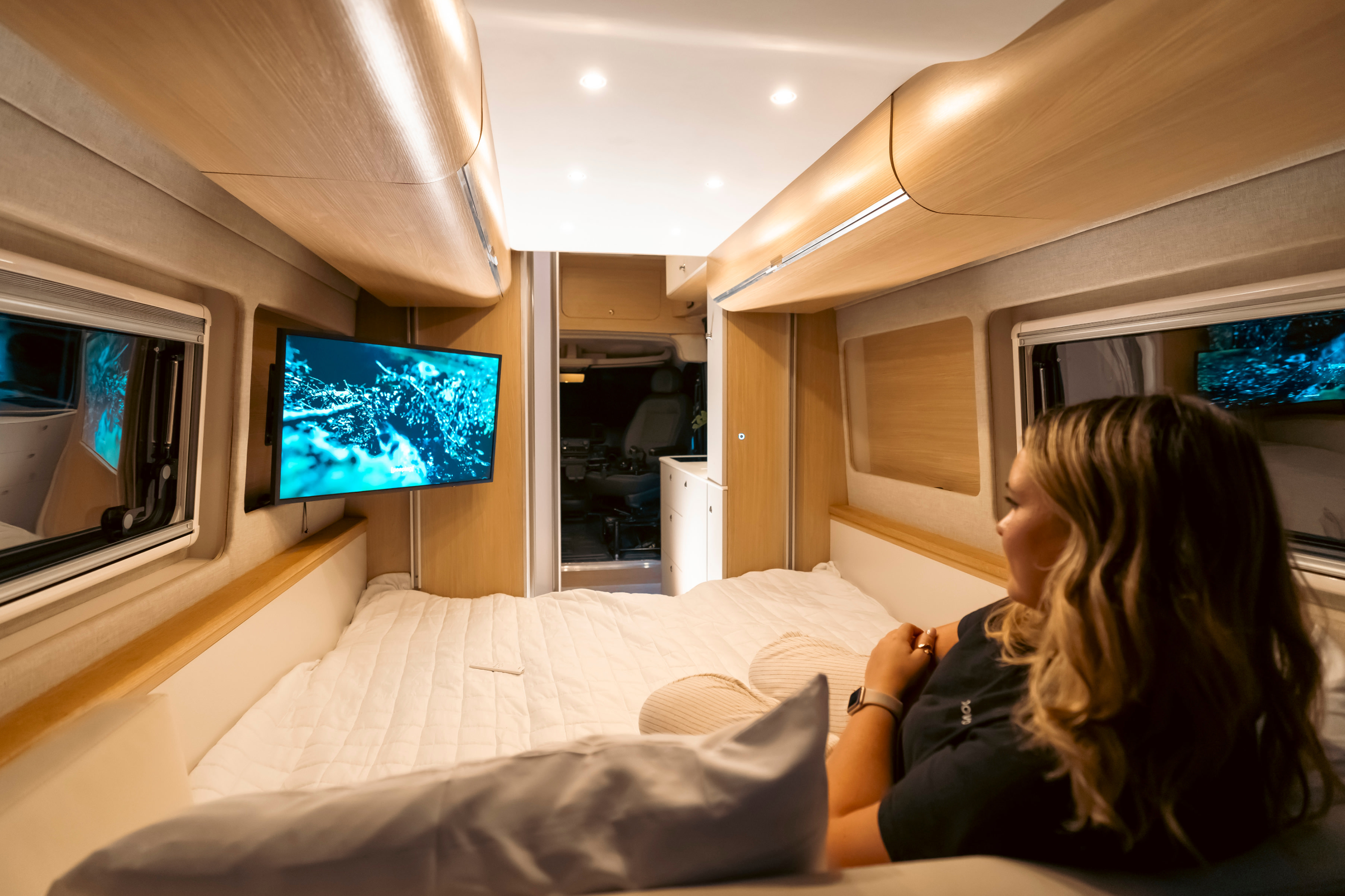 A woman watches TV in a well-lit, modern RV rental bedroom, offering comfort and entertainment on the road.. Noovo Plus 2024