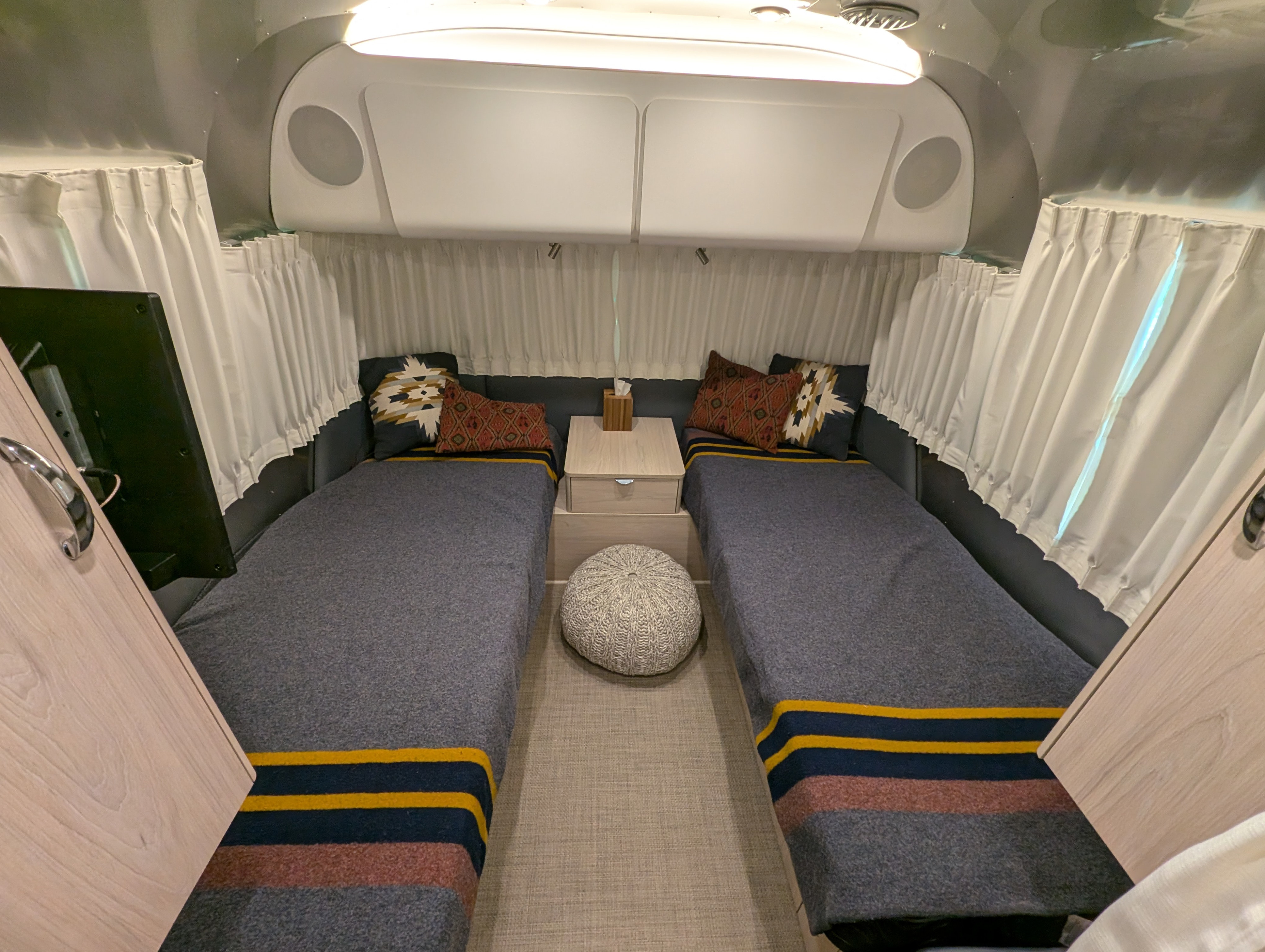 Inside a modern **RV rental**: twin beds, central nightstand, white curtains, and storage.. Airstream Globetrotter 2024