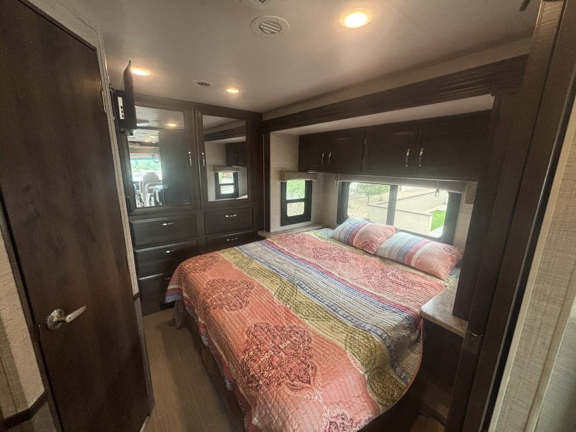 Cozy RV rental bedroom with a colorful quilt, ample dark wood storage, and bright windows, offering comfortable travel.. Entegra Coach Vision 2021