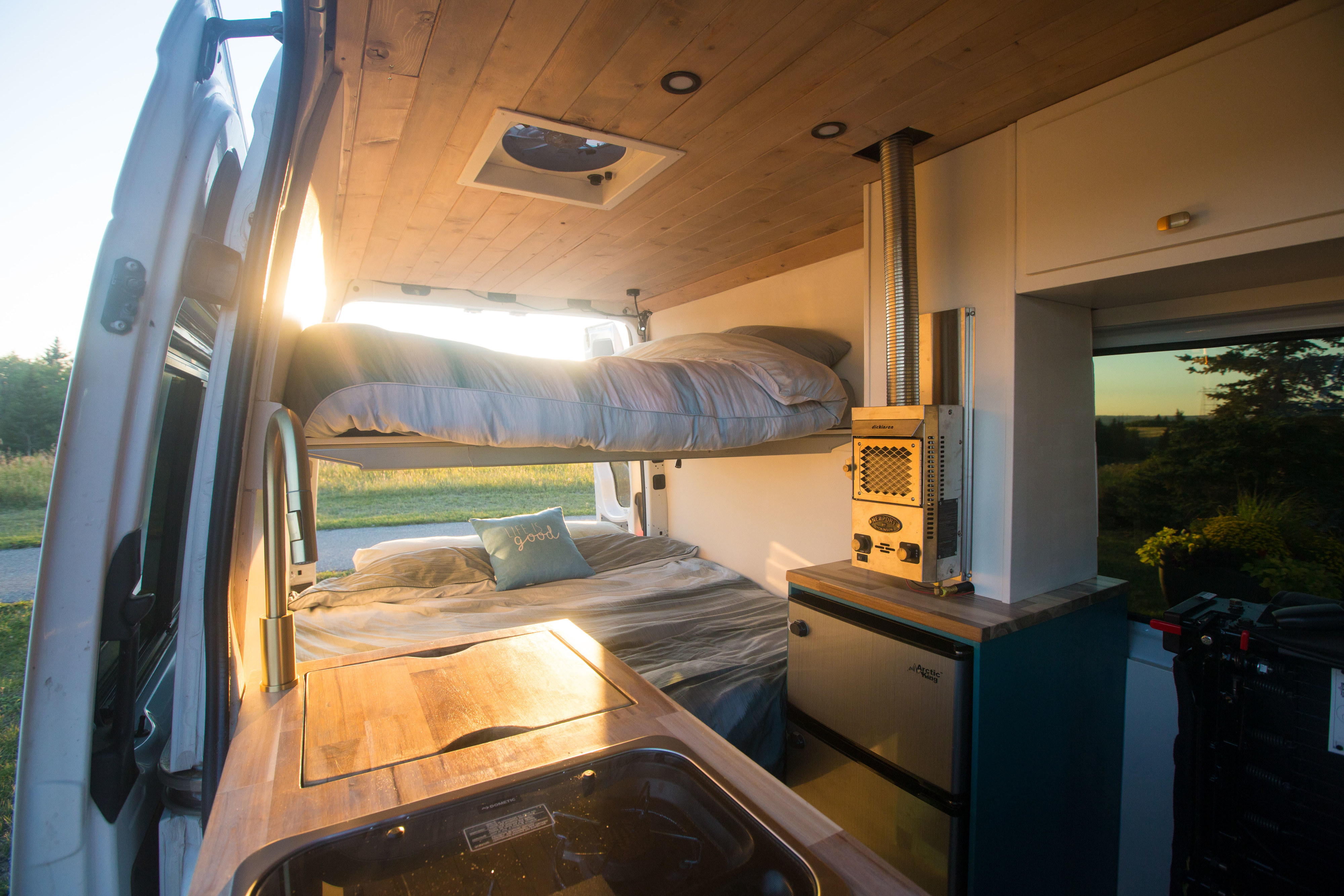 Cozy RV rental interior with bunk beds, kitchen sink, heater, and fan, bathed in warm sunset light from open doors.. Mercedes-Benz Sprinter 2015
