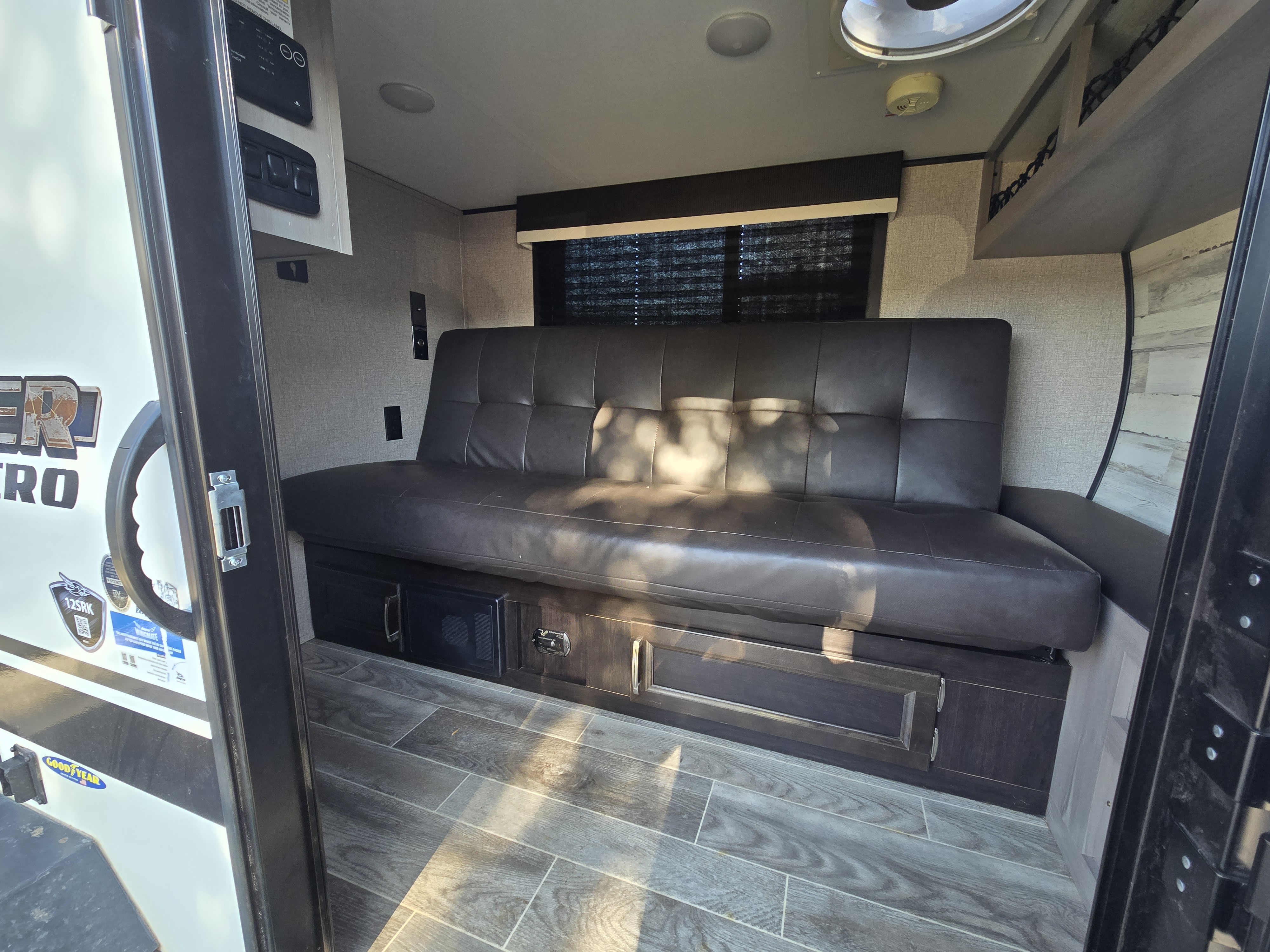 Inside an RV rental: a dark brown futon sofa with storage, wood-look flooring, and wall controls, ready for travel.. Jayco Jay Feather Micro 2021