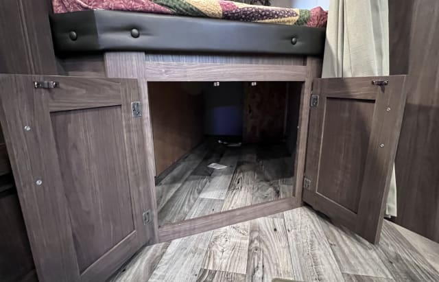 Open storage doors under an RV rental bunk bed, revealing a spacious compartment with wood-look flooring & interior lighting.. Keystone RV Hideout LHS 2019