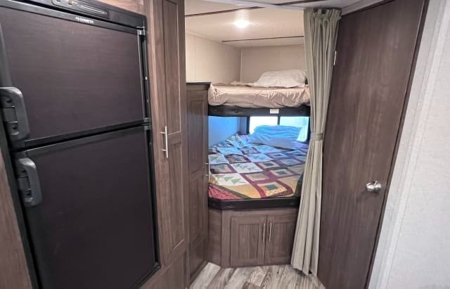 Inside an RV rental: view of a large refrigerator, wood-paneled storage, and cozy bunk beds with colorful bedding.. Keystone RV Hideout LHS 2019