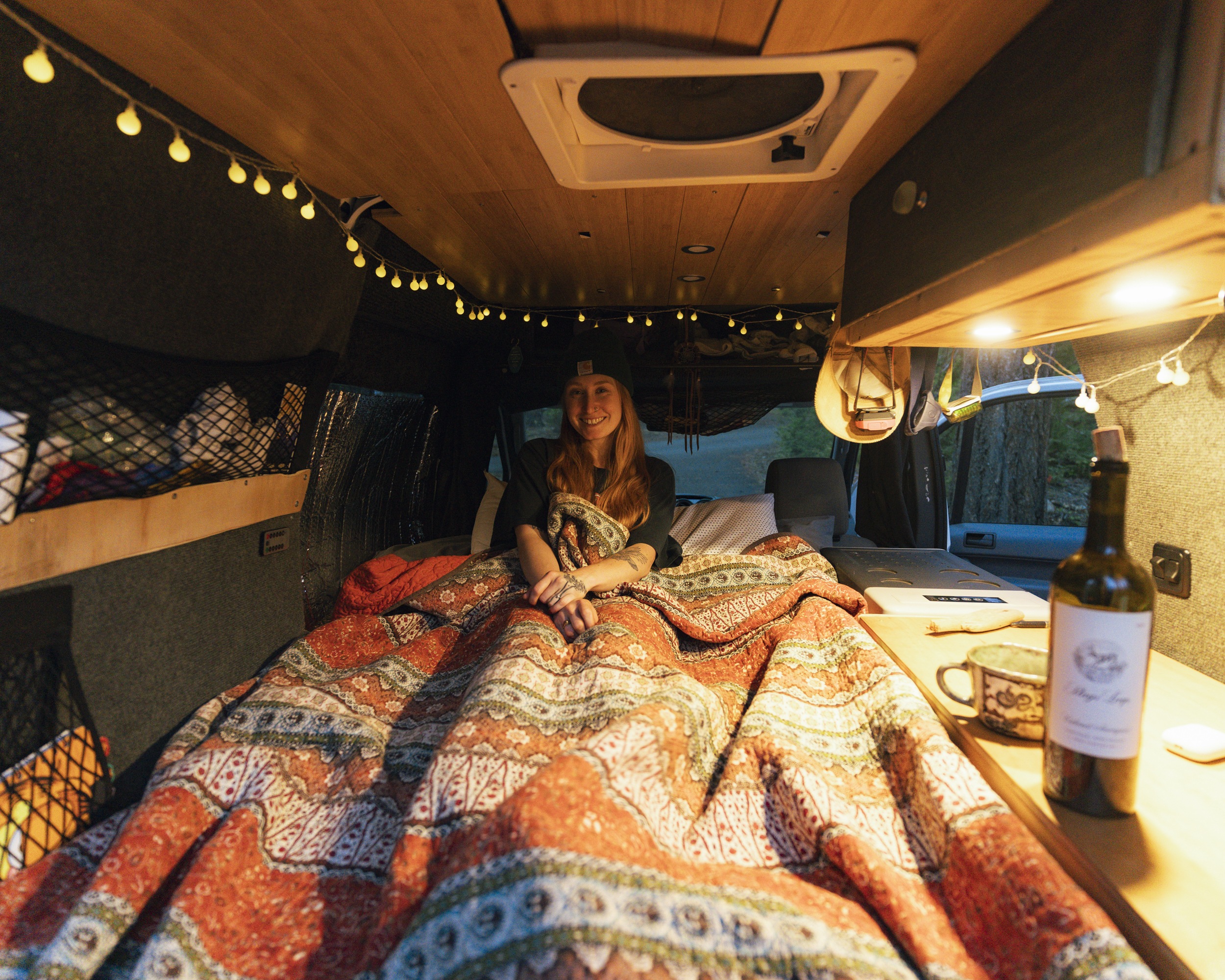 A woman smiles cozily in an RV rental, covered by a patterned blanket with warm string lights above.. Ford Transit 2011