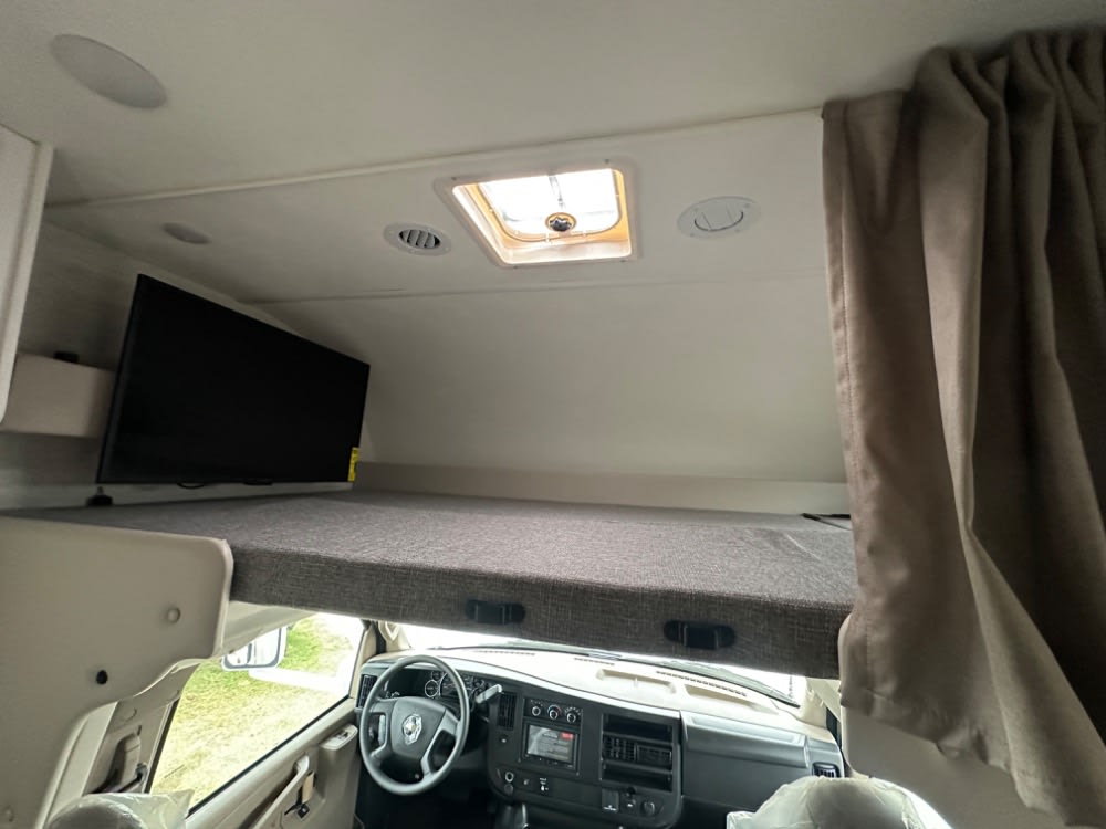 Inside an RV rental: a comfortable cab-over bunk with TV, ceiling lights, and driver's area below.. Thor Freedom Elite 2024