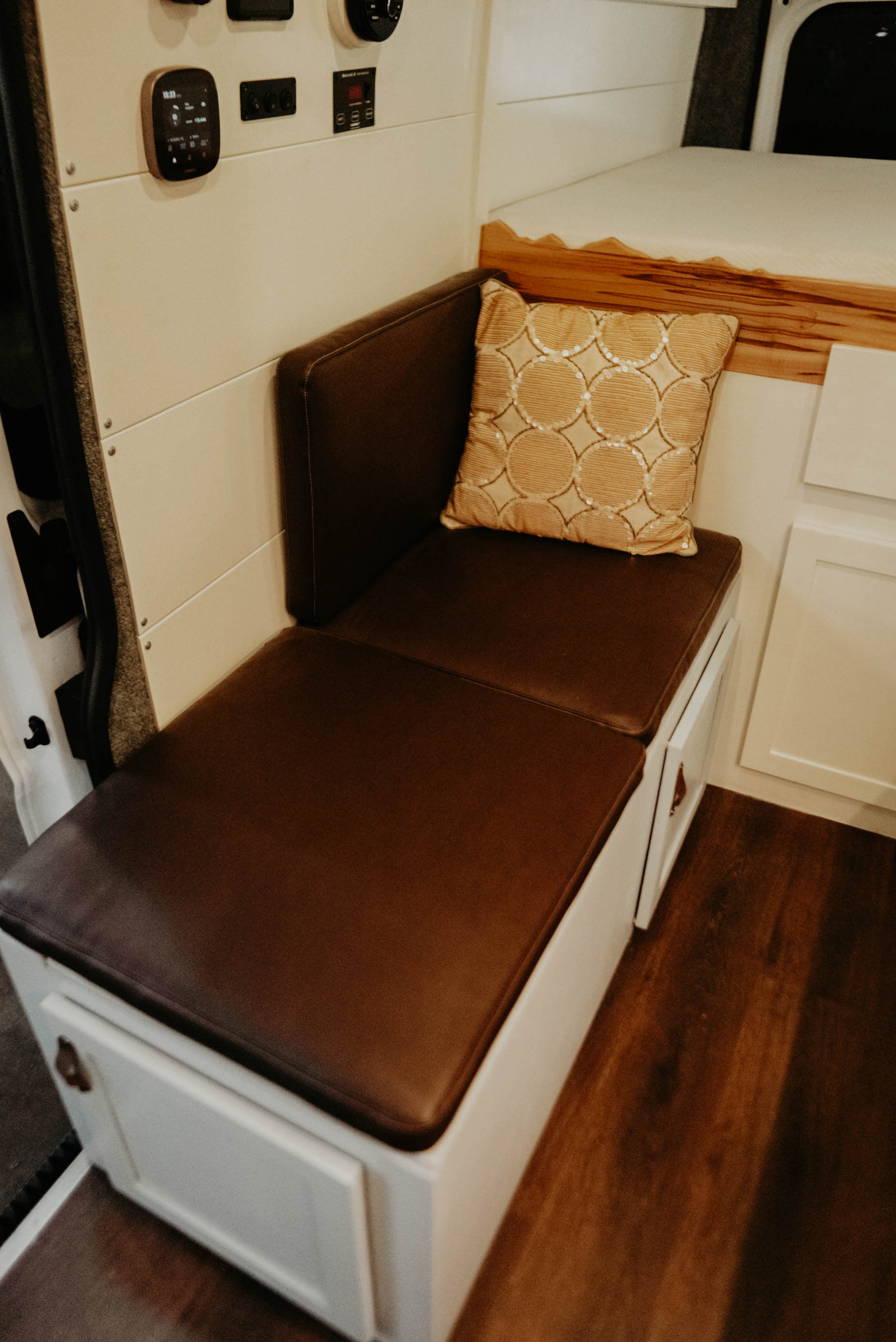 Cozy RV rental interior: brown cushioned bench with storage, patterned pillow, a raised bed, and wall controls.. Noke Van Co. Rory 2021