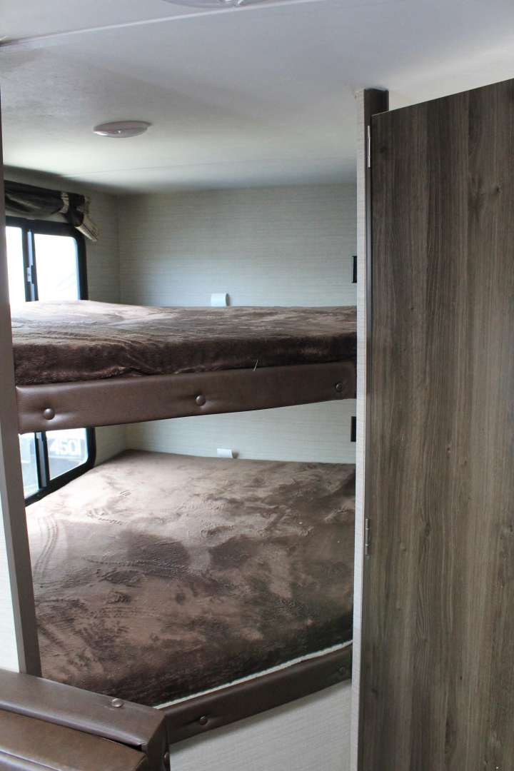 RV rental interior: Stacked bunk beds with brown blankets, window view, and wooden door. A compact sleeping area.. Keystone RV Bullet Crossfire 2019