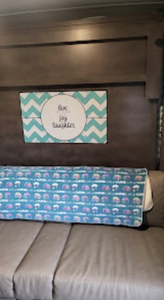 Inside an RV rental: a brown couch, a blue blanket with small campers, and a "live joy laughter" sign on the wooden wall.. Forest River Rockwood 2021